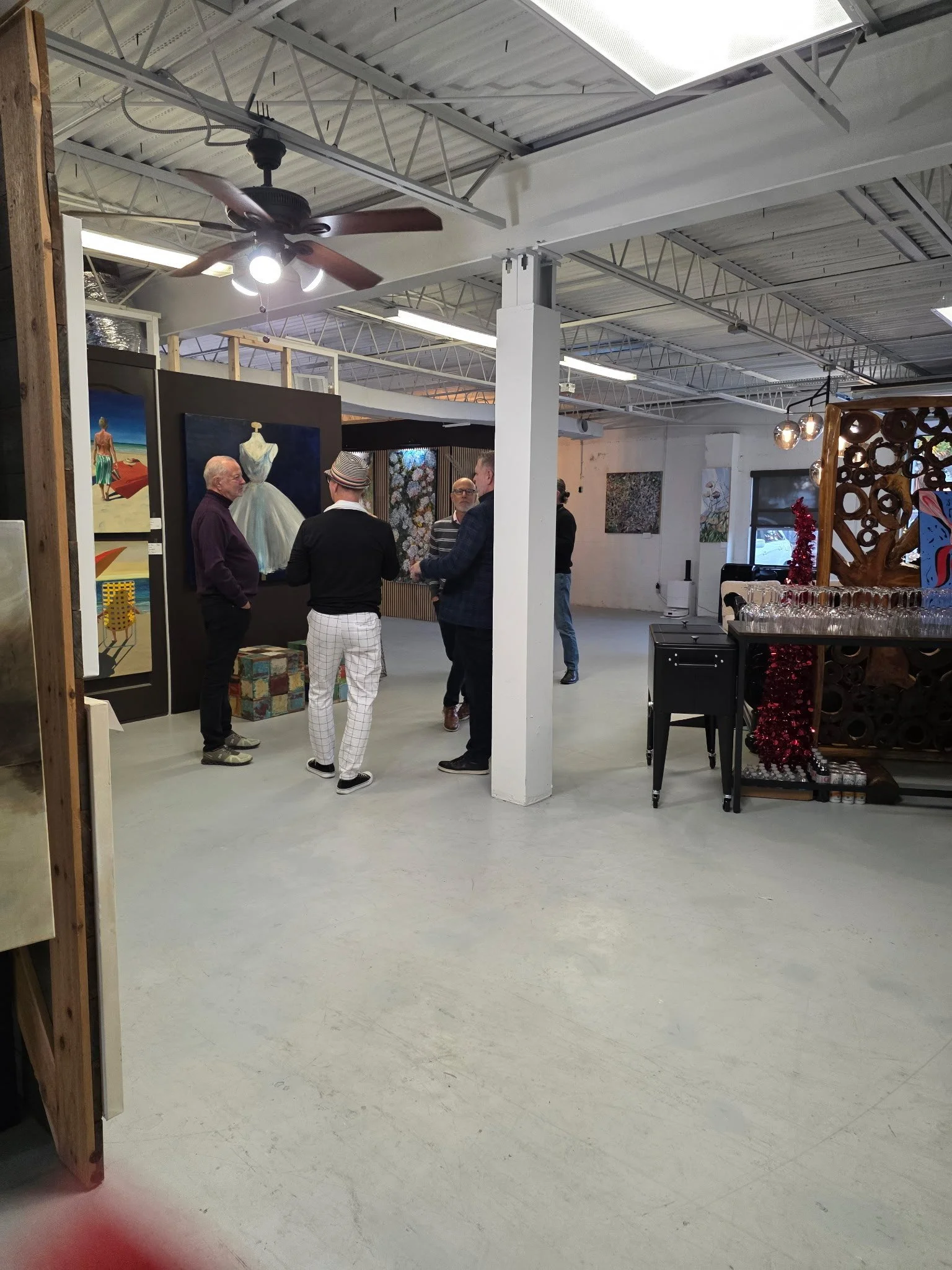 Group of people mingling in an art gallery with paintings on the walls and sculptures, a ceiling fan overhead, and various decorative items.