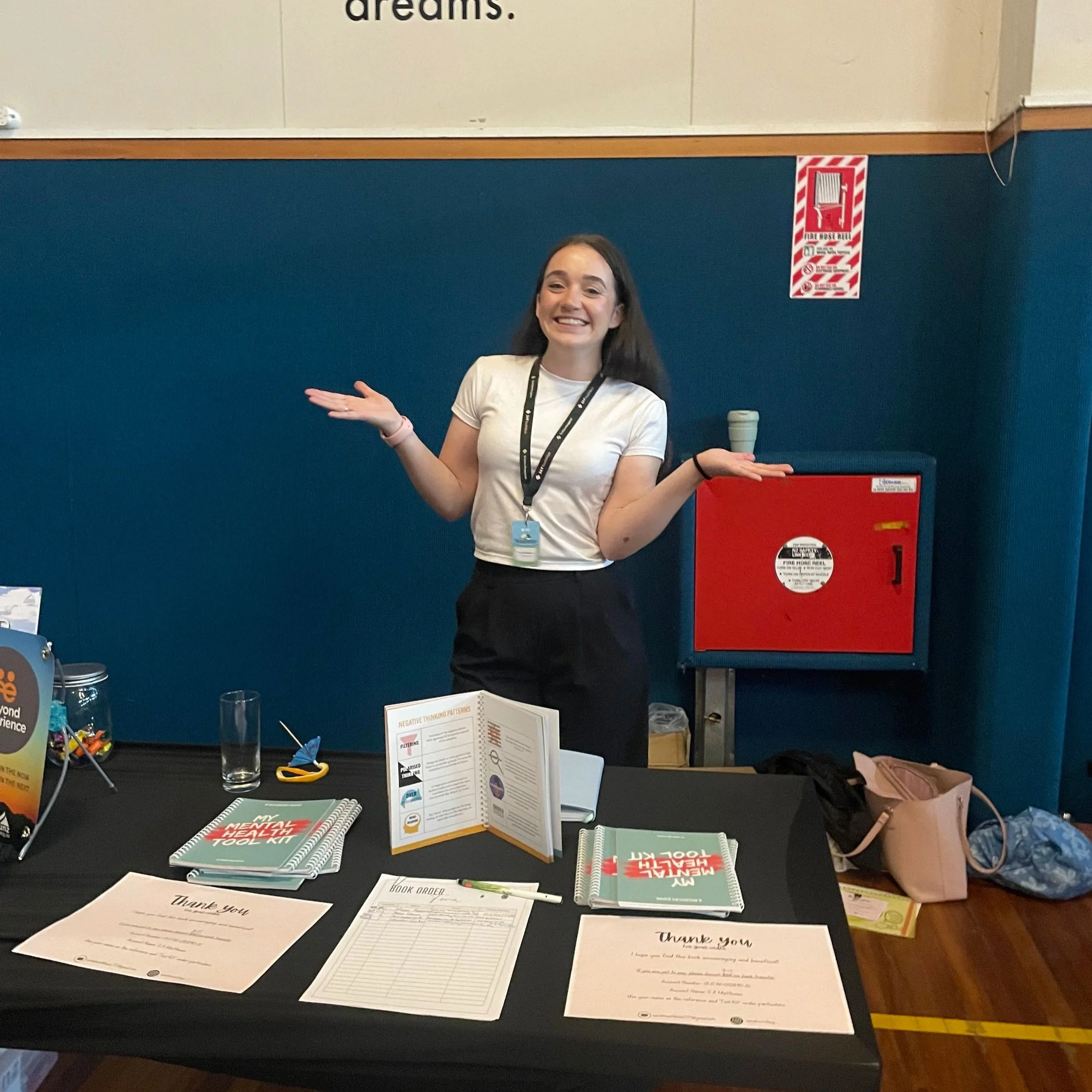 Sarah Matthews, a young woman, standing behind a table at a conference, exhibiting and selling her book, My Mental Health Tool Kit: A Recovery Guide, happy and smiling with arms outstretched.