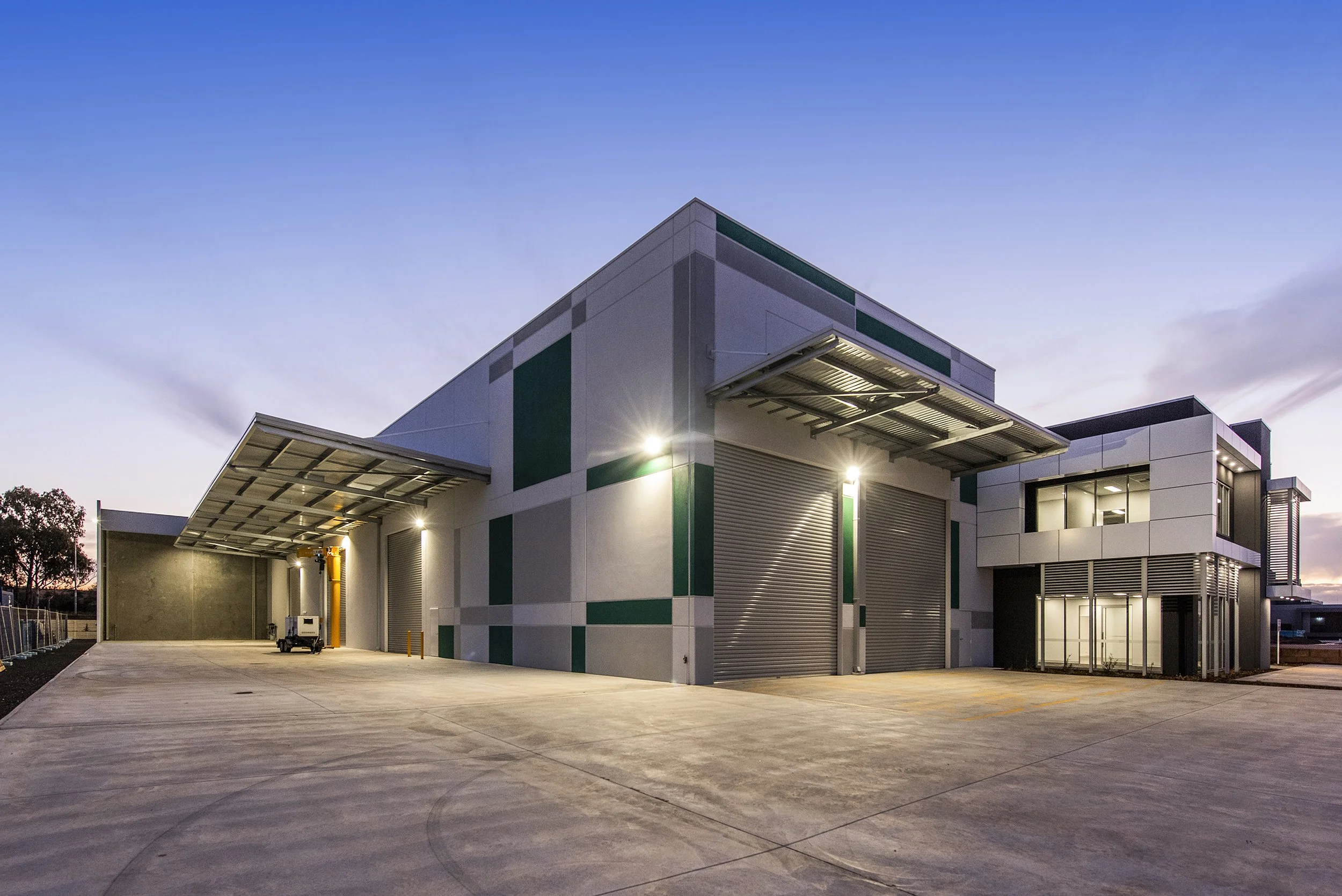 Modern industrial warehouse building at dusk with large security shutters and metal awnings, surrounded by concrete pavement.