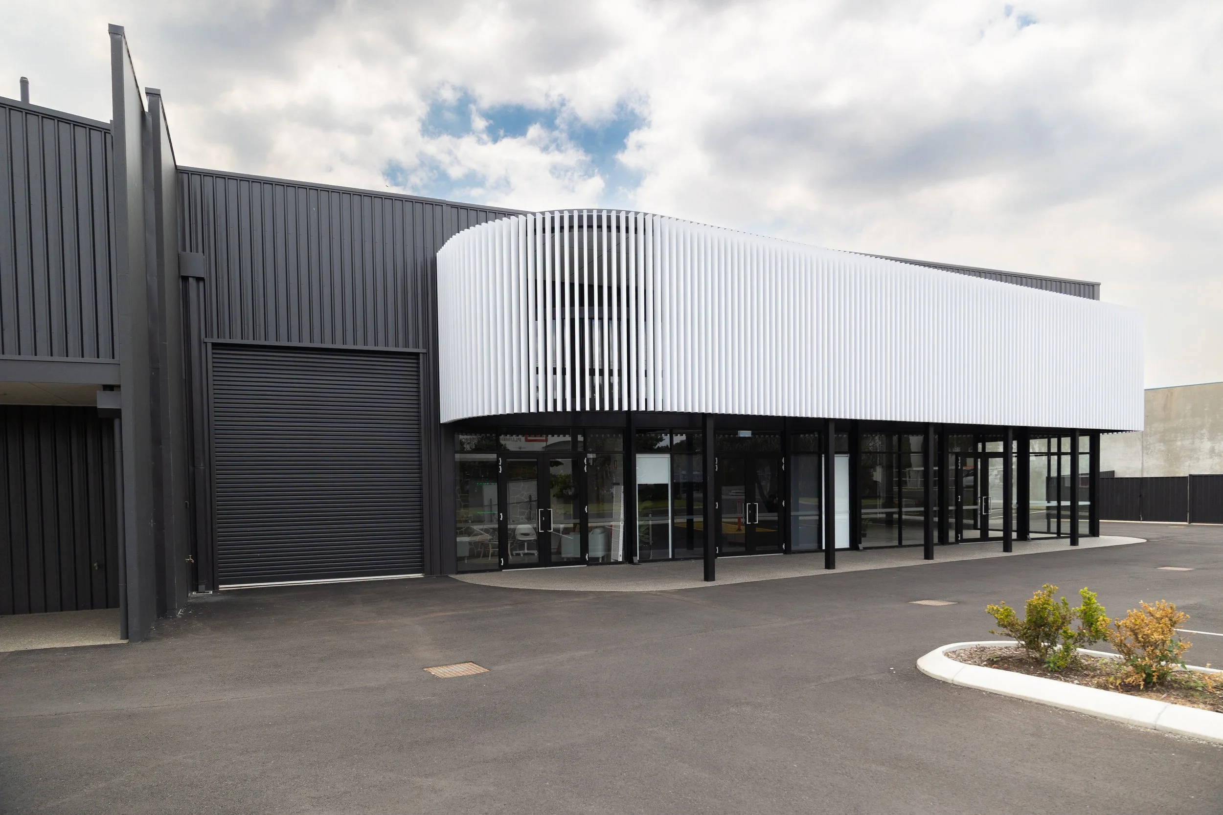 Modern commercial building with black exterior walls, large glass windows, and a white curved facade with vertical slats on the upper level. There are small bushes and a parking lot in front.