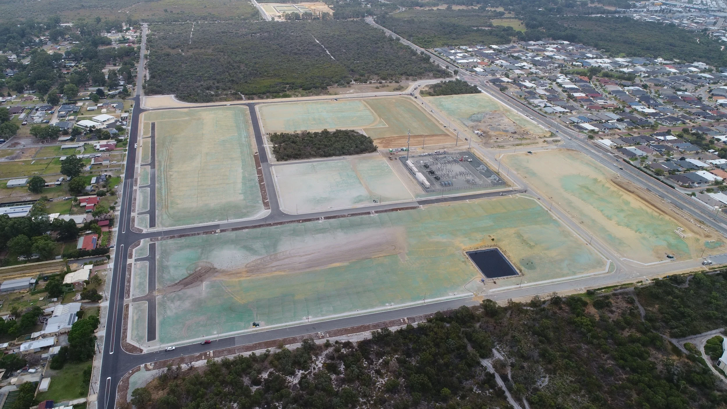 Aerial view of an area under development with empty lots, roads, and some structures, surrounded by residential houses and trees.