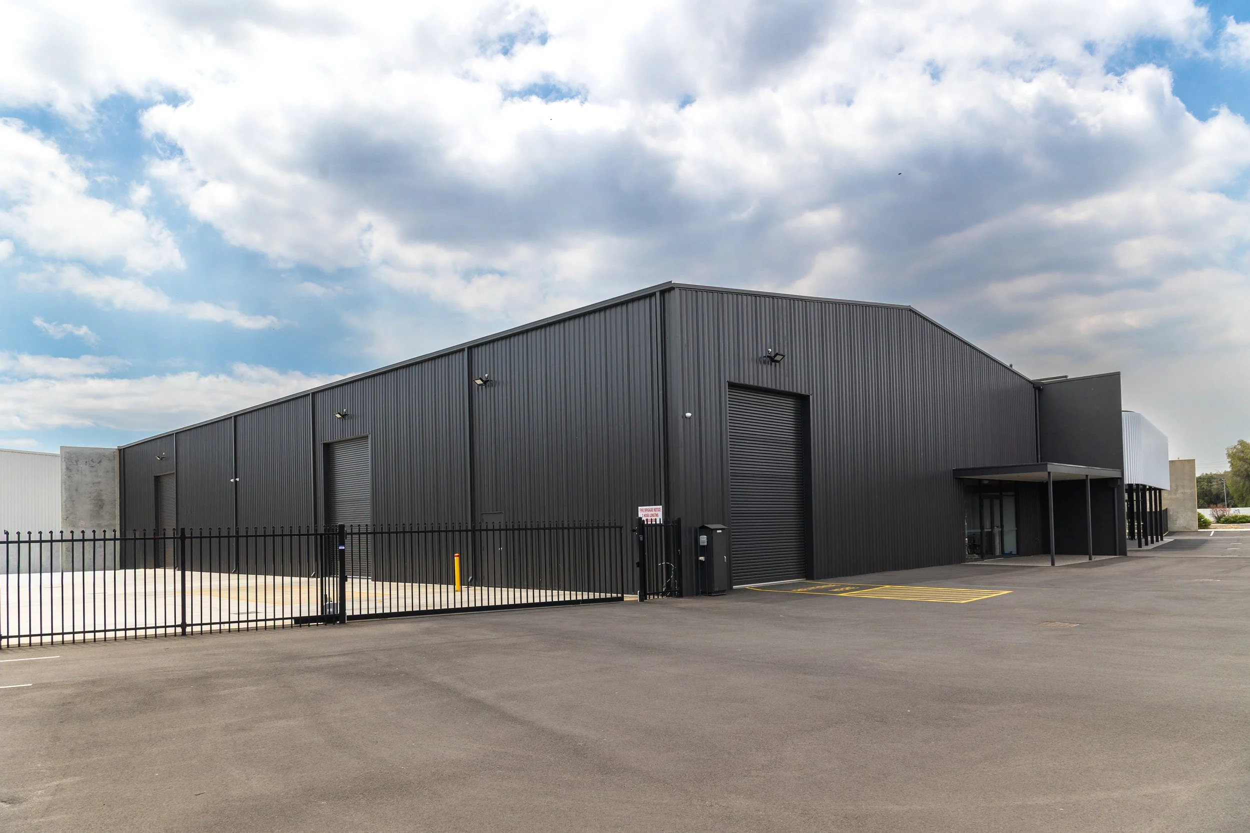 A large industrial warehouse with black metal siding, multiple closed large roll-up doors, a small covered entryway, and surrounding parking lot under a partly cloudy sky.