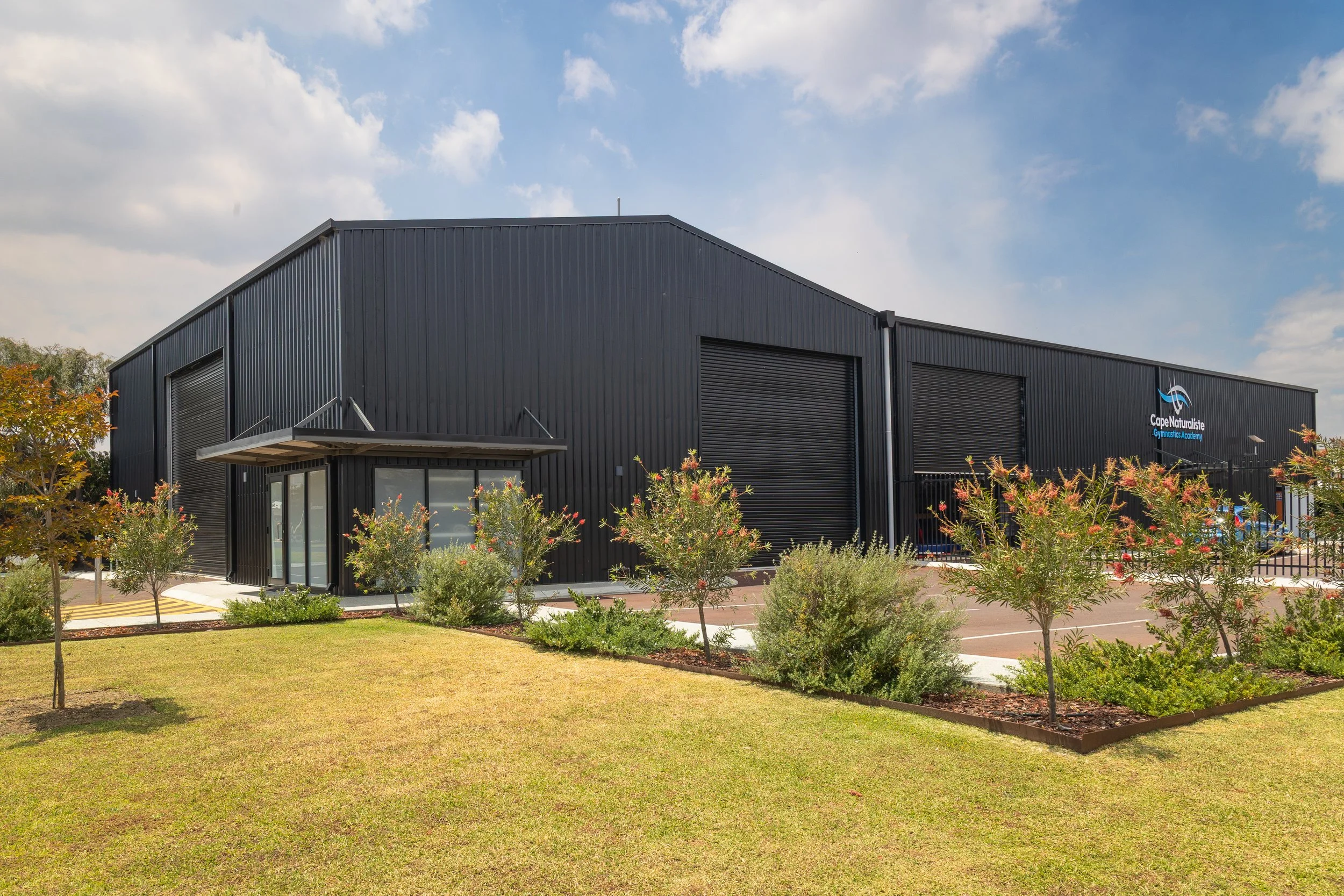 A large black industrial building with a sign that reads 'Cape Naturaliste Gymnastics Academy,' surrounded by a well-kept lawn and small trees, under a partly cloudy sky.