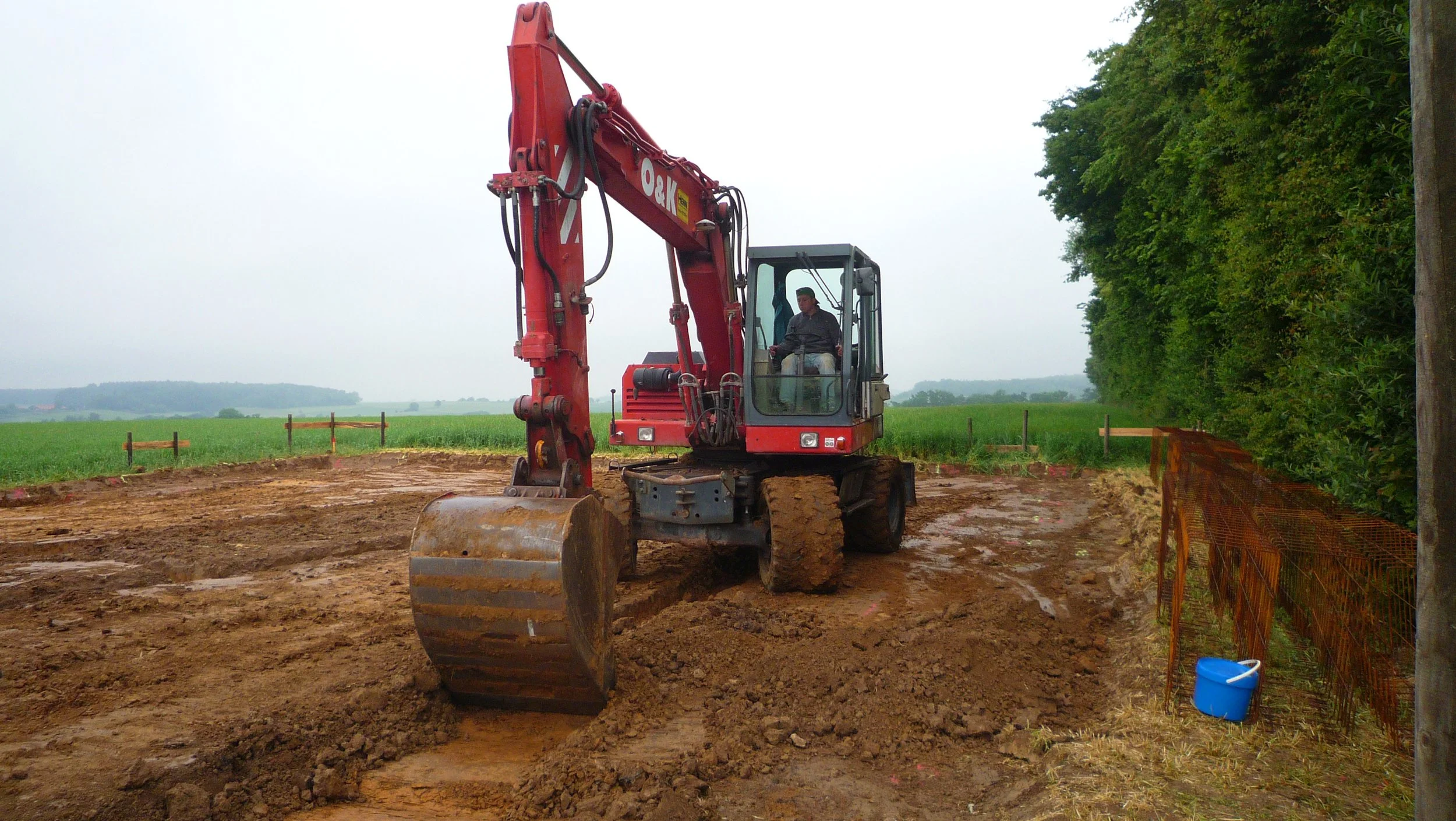 A person operating a red excavator on a construction site in a rural area, with dirt and a green field in the background, and a row of trees on the right.