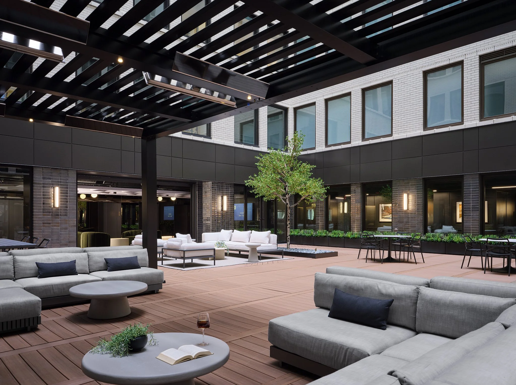 Modern outdoor patio with gray sofas, round tables, a tree in the center, and black dining tables with chairs, surrounded by a glass-walled building. Building Amenities, workplace interiors, hospitality, office, interior design, interiors, downtown