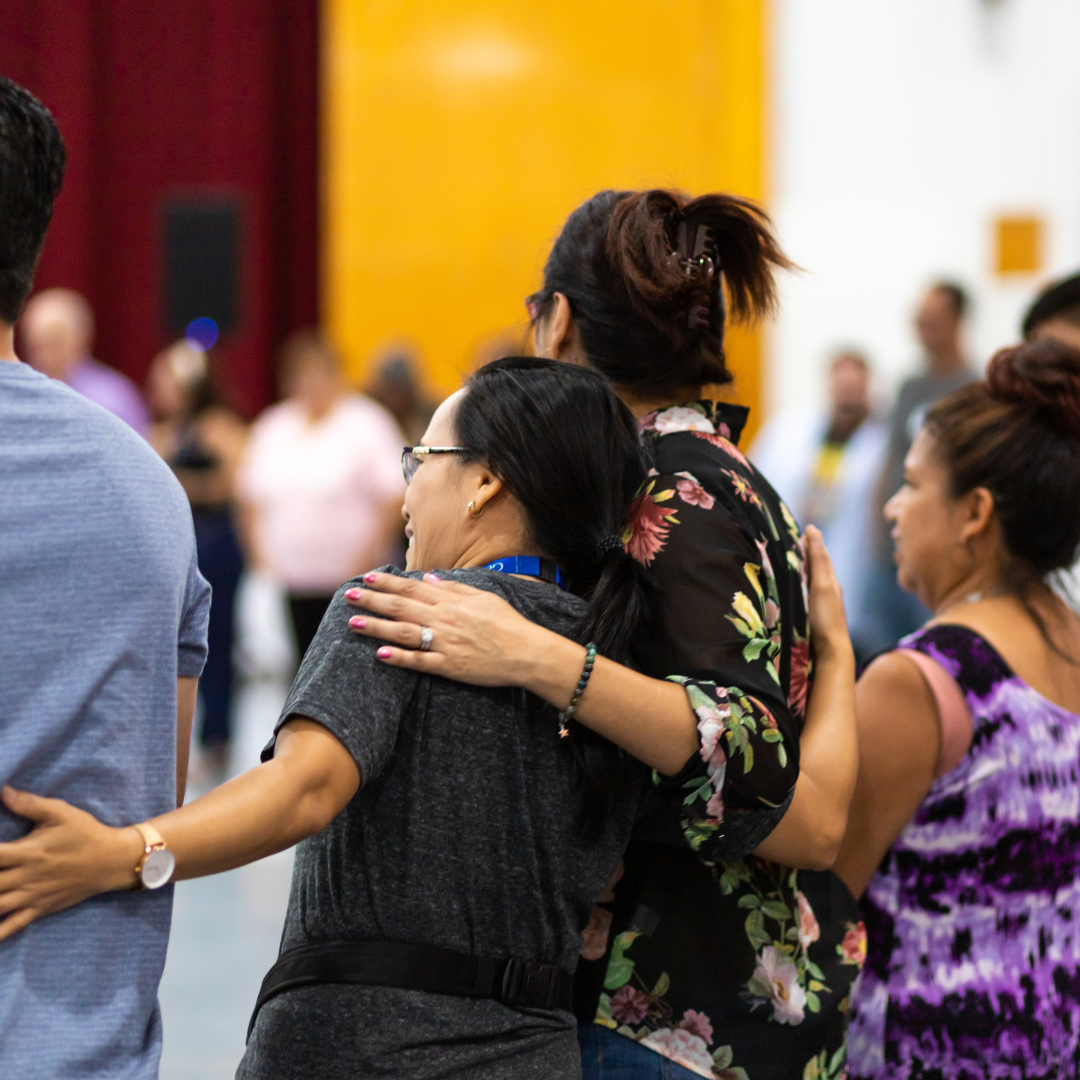 Two women embracing at a gathering, surrounded by other people, in a room with colorful walls.