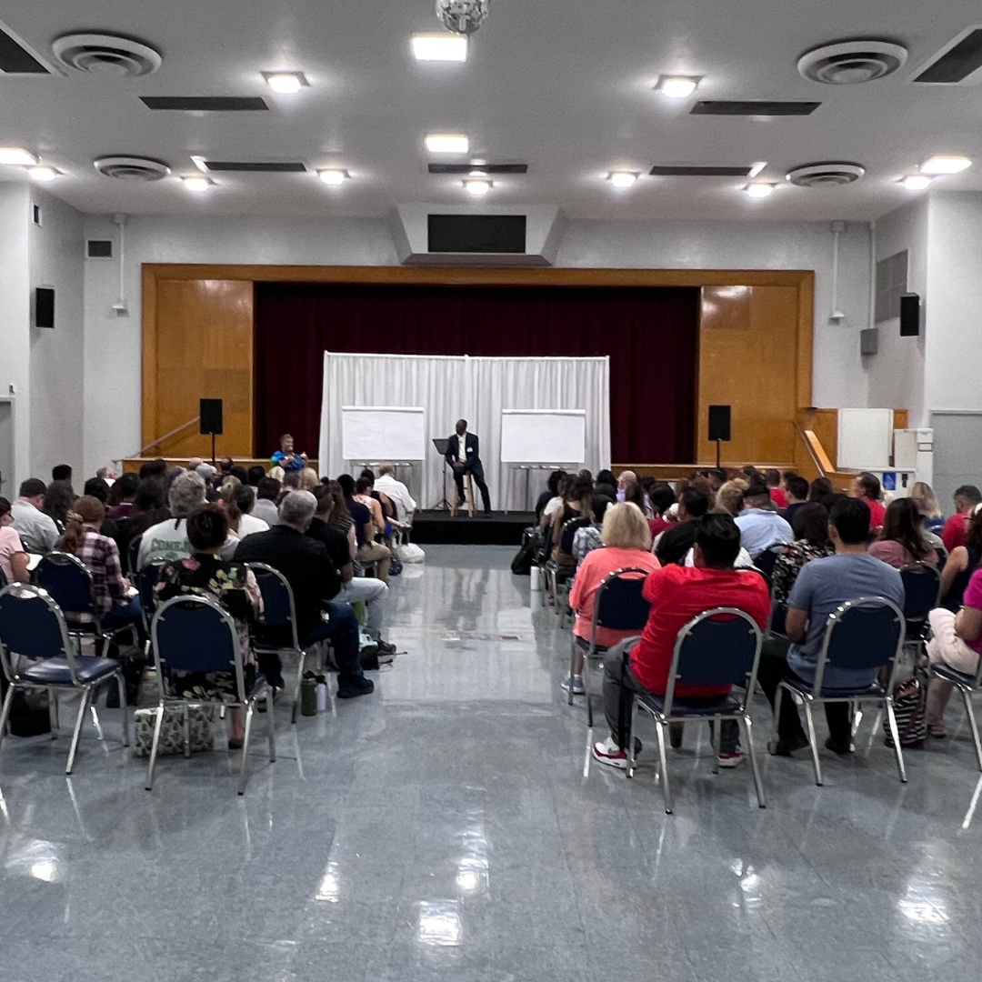 Indoor conference or seminar with a large audience listening to a speaker on stage, who is seated with two whiteboards behind him.