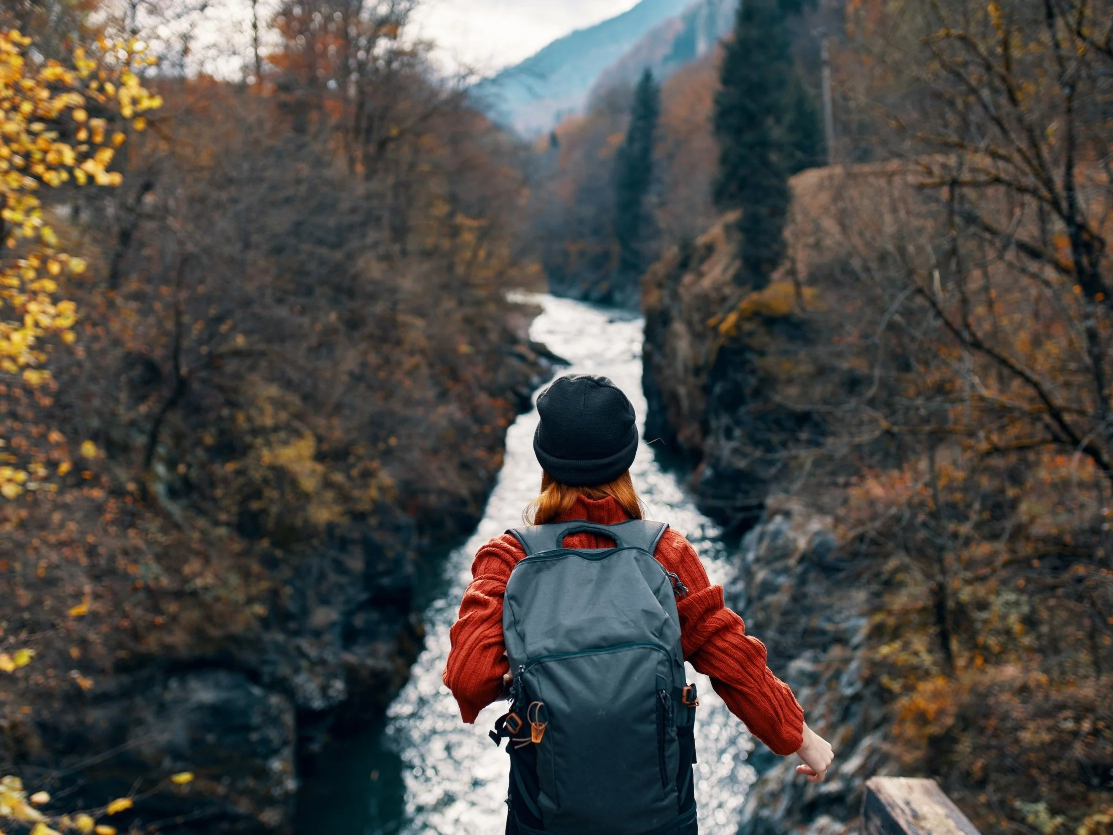 A person with red hair wearing a black beanie, red jacket, and gray backpack stands overlooking a canyon with a river running through it, surrounded by autumn-colored trees.