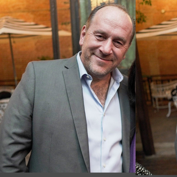 A middle-aged man with a receding hairline, beard, and mustache, wearing a light blue dress shirt and gray blazer, smiling at the camera in an outdoor setting at sunset with wooden structures in the background.
