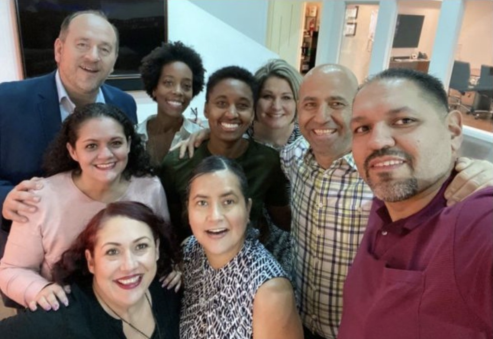 Group of nine diverse people smiling and gathered together for a photo indoors, some with arms around each other, in a friendly, casual setting.
