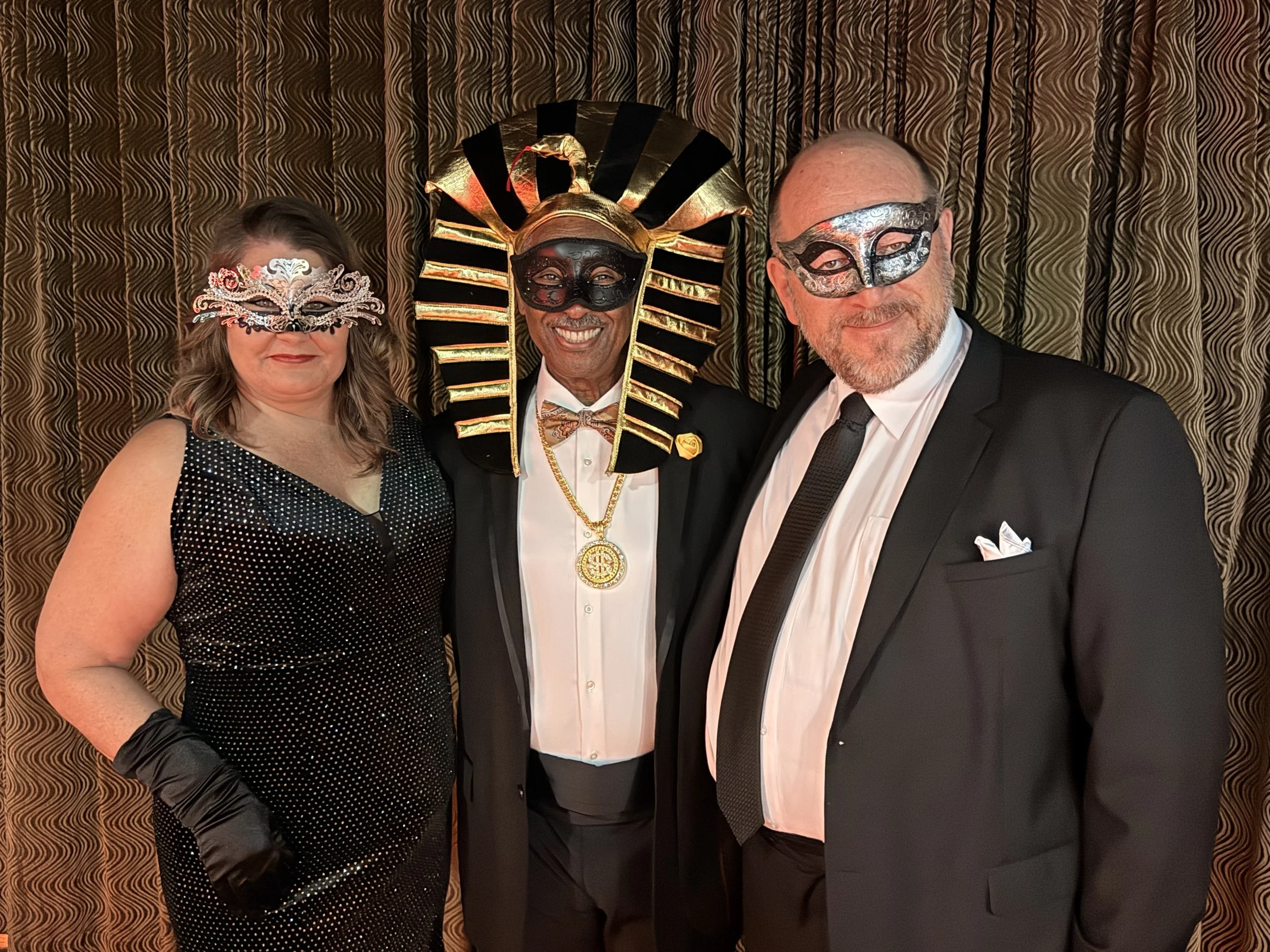 Three people wearing fancy masks and costumes at a Mardi Gras or masquerade event, standing in front of a patterned curtain.