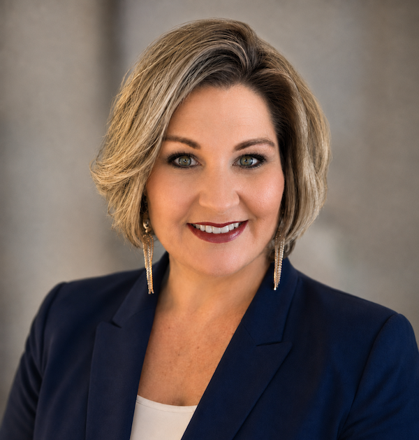 Kristen Bowers, coach, wearing a navy blazer, cream blouse, blue necklace, and a smartwatch sitting at a table in a cafe with a window behind her showing a tree and cars outside, and floral wall decorations in the background.