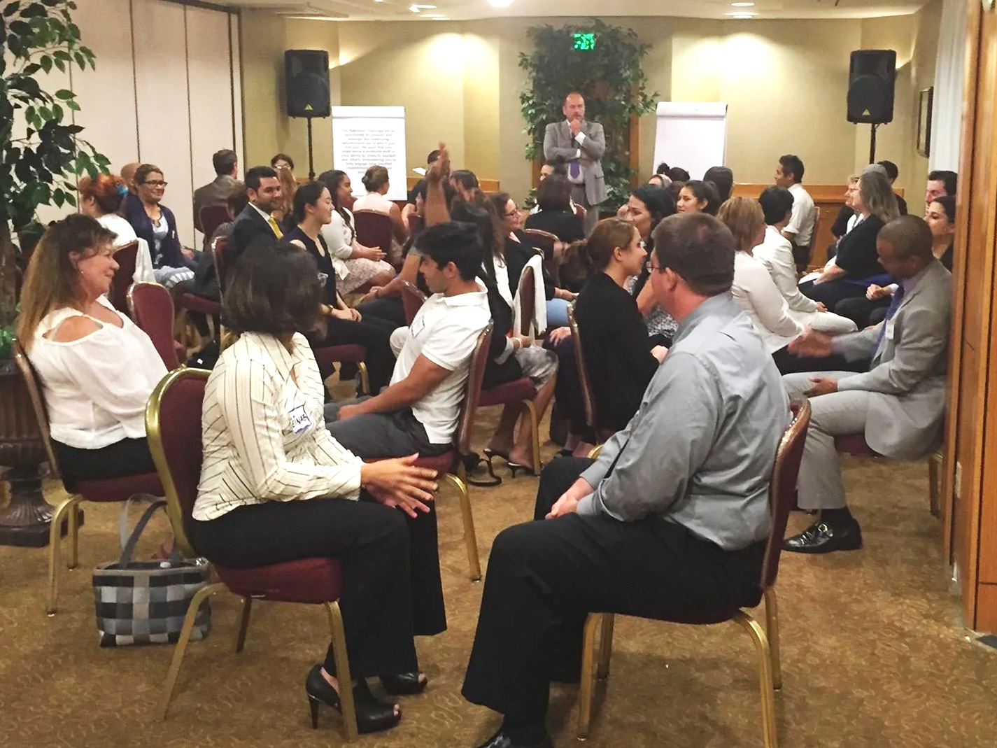 A group of people sitting on chairs in a conference room, facing a speaker at the front who is standing and speaking into a microphone, with a whiteboard and a potted plant behind him.