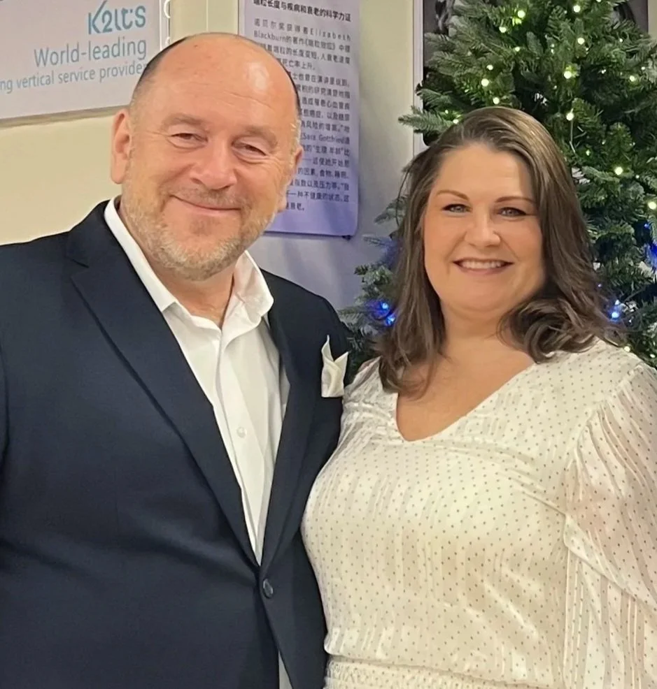 A man in a dark suit and a woman in a light dress standing in front of a Christmas tree decorated with lights.