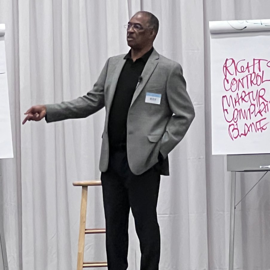 Man in gray blazer giving a presentation, pointing to the left, standing in front of a whiteboard with handwritten notes.