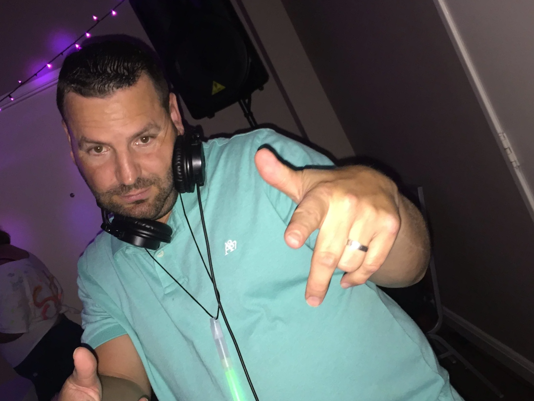 A man wearing a light blue polo shirt, with headphones around his neck, points at the camera with both hands, giving a thumbs-up gesture. Purple lights are visible in the background.