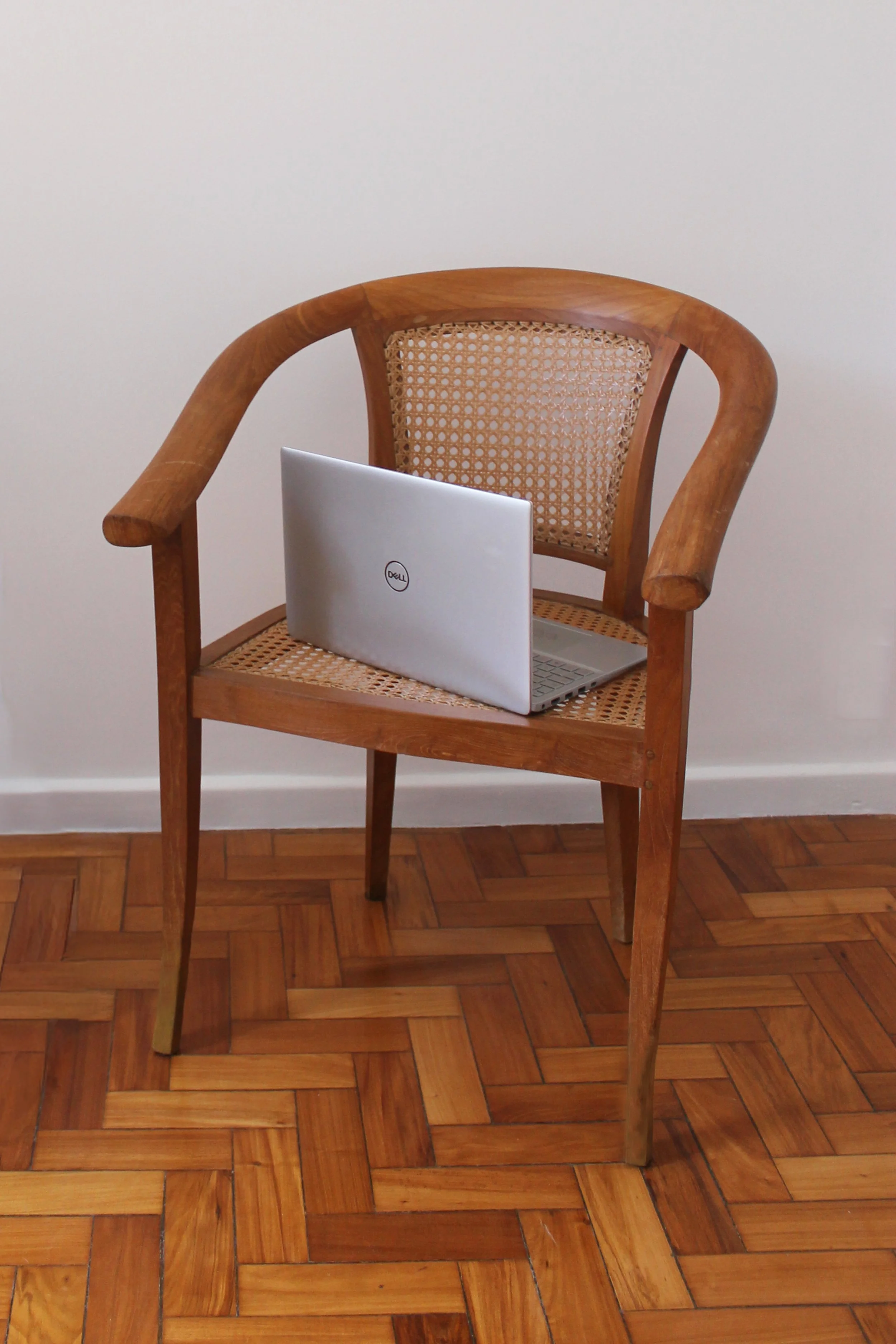 Cadeira de madeira com encosto de cana e braço arredondado, com um laptop Dell na parte da frente, sobre o assento, em uma sala com piso de parquet e parede branca ao fundo.