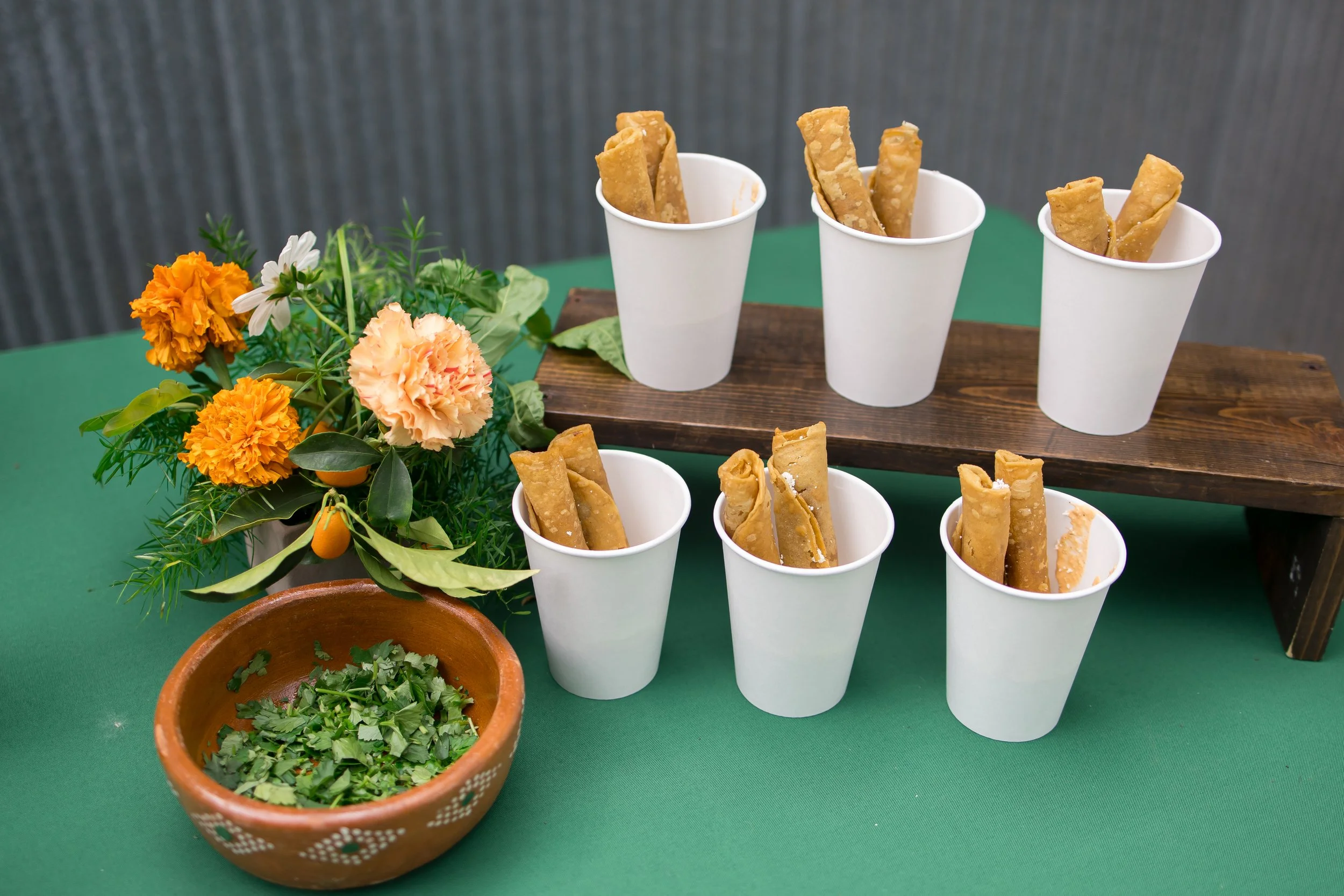 A display of fried snack rolls in white cups on a wooden stand, with colorful flowers and green herbs in a terracotta bowl on a green table.