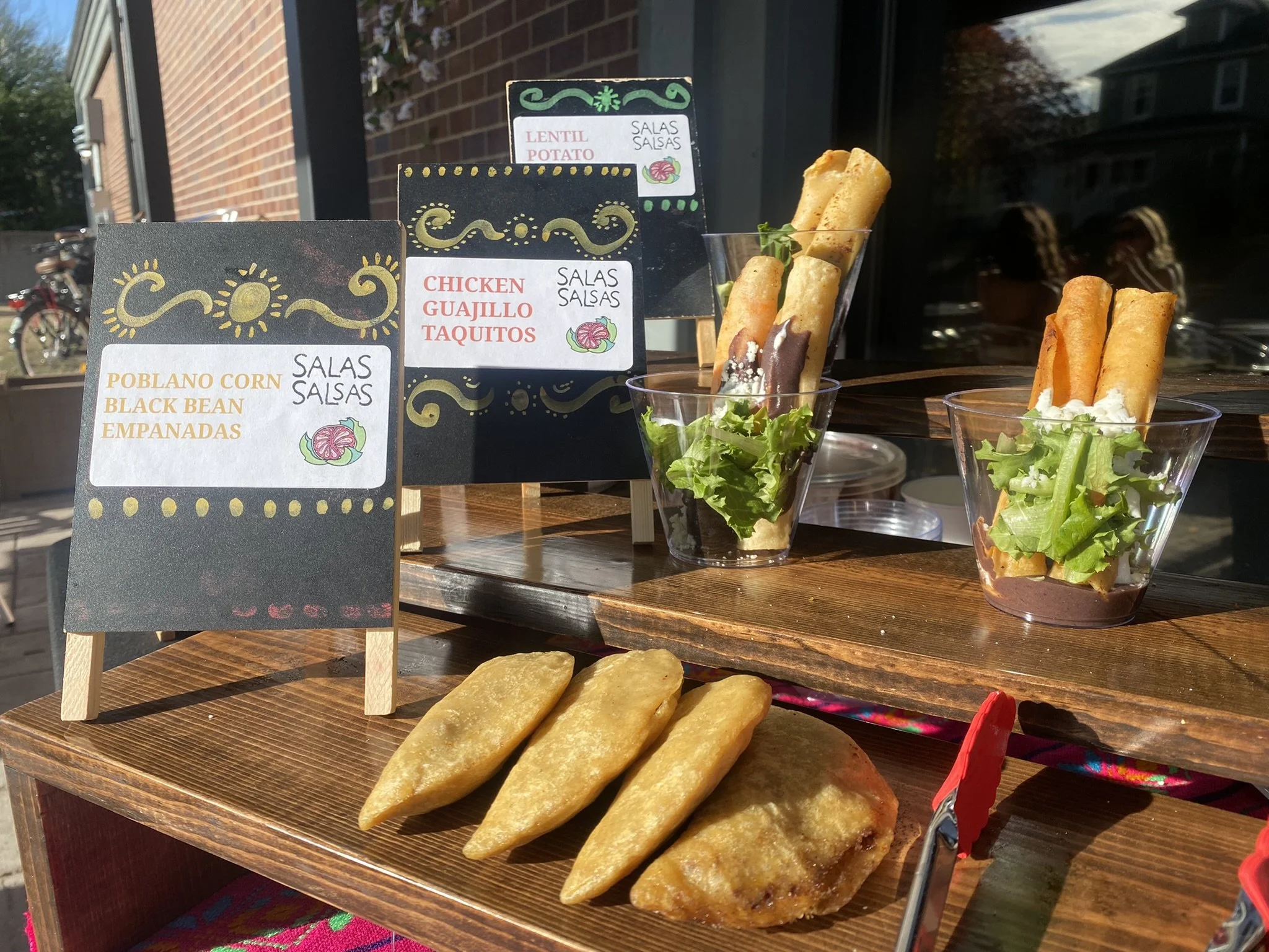 Display of Mexican food items including taquitos with lettuce, cheese, and sauces, along with black bean empanadas and crispy breadsticks, placed on a wooden serving tray with small chalkboard signs describing the dishes.
