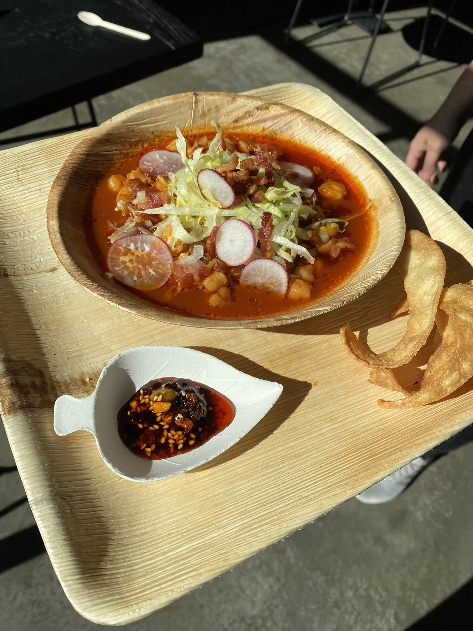 A wooden tray holding a bowl of spicy soup garnished with radish slices, shredded lettuce, and bacon bits, with a side of potato chips and a small dish of spicy chili sauce.