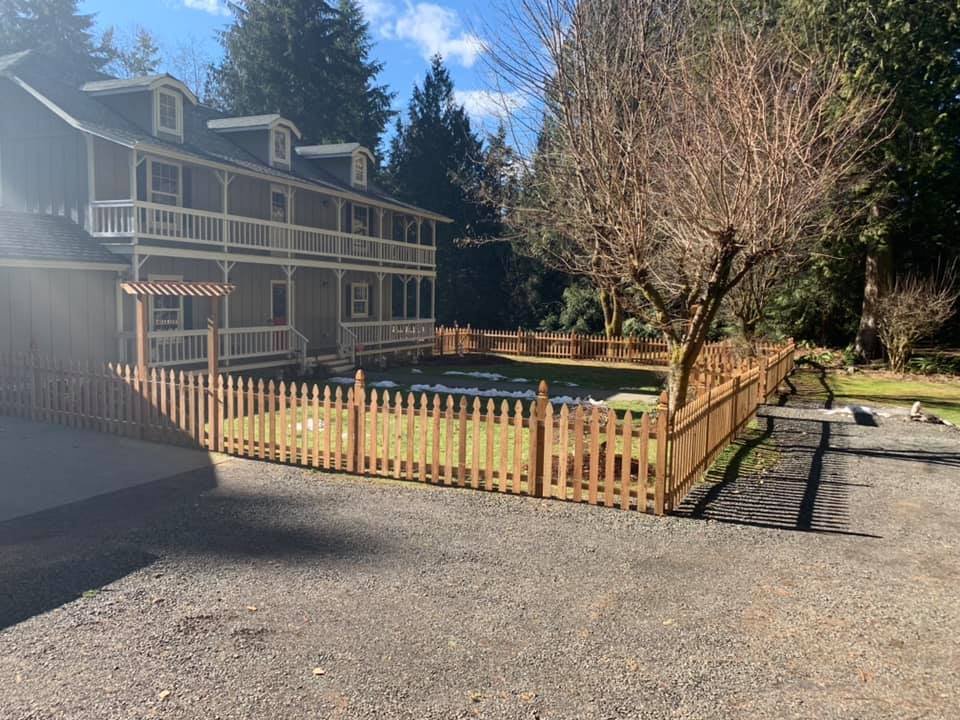 A fenced-in yard with a leafless tree, green grass, and a treeline in the background, in front of a gray residential building with multiple balconies.