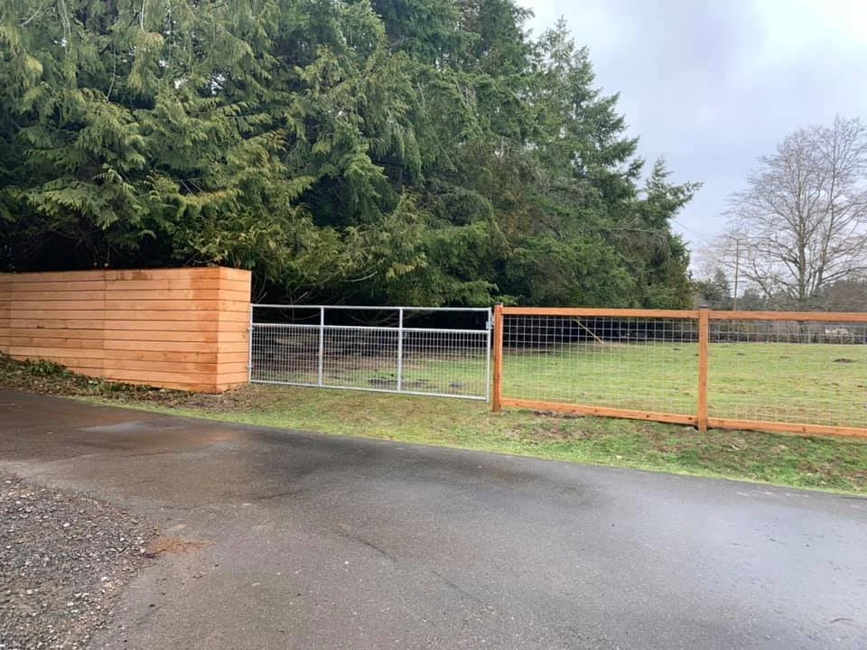 A metal gate and fence on a grassy area next to a paved road, with a large tree and a wooden fence on the left side.