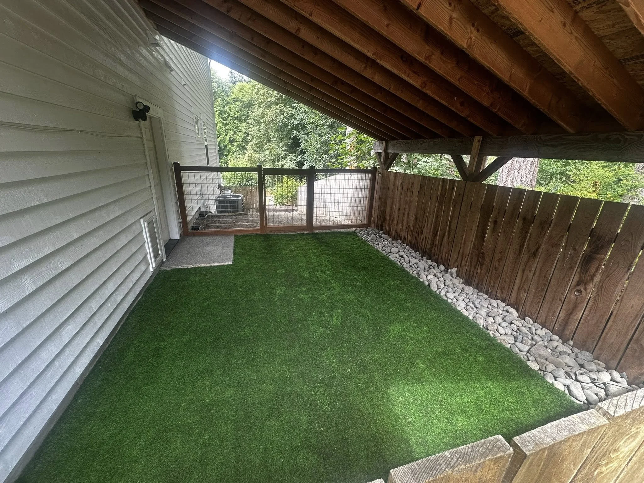 A small backyard area with artificial grass, a gravel border, a wooden privacy fence, and a house wall on the left, partially covered by a wooden roof.