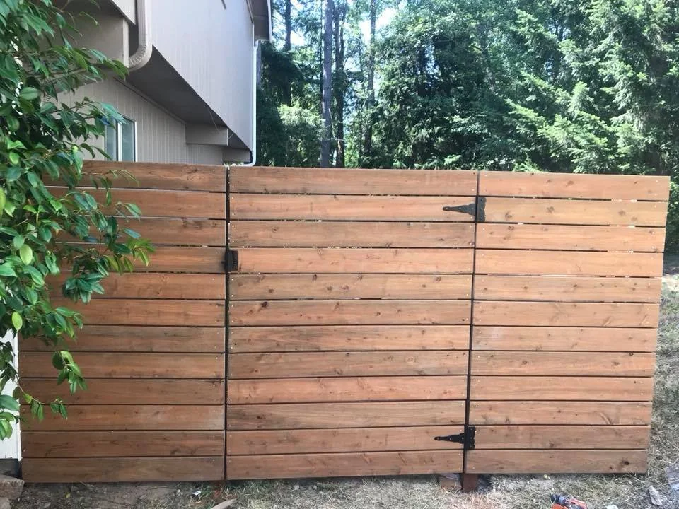 New wooden privacy fence with hinges and latch, partially open, in a backyard with trees and plant greenery.
