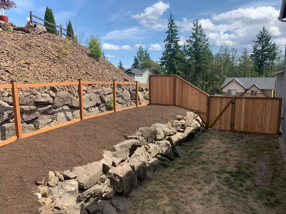 A backyard with a newly built wooden fence, a stone retaining wall, and a dirt slope. Trees and residential houses are in the background under a partly cloudy sky.