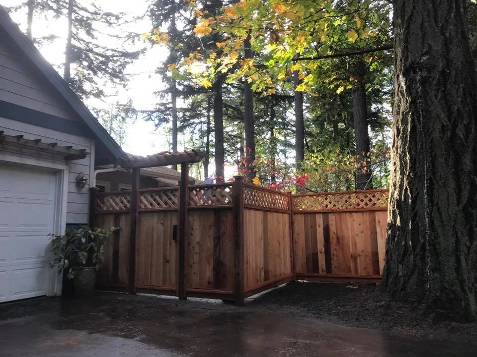 A newly-installed wooden privacy fence surrounds a backyard area next to a house with a white garage door, large trees with autumn leaves are visible in the background.