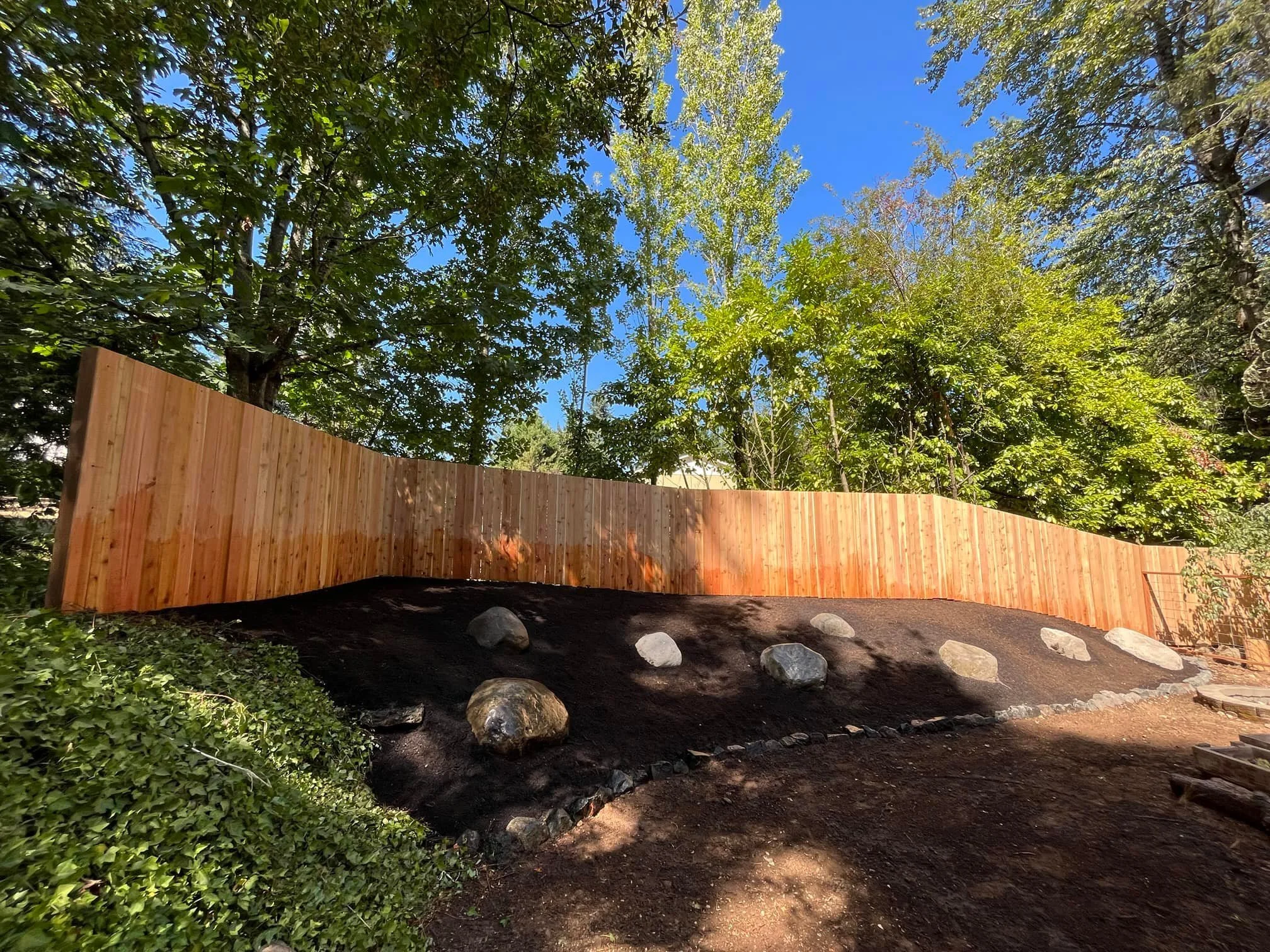 Wooden privacy fence in a garden with trees, rocks, and a dirt pathway.
