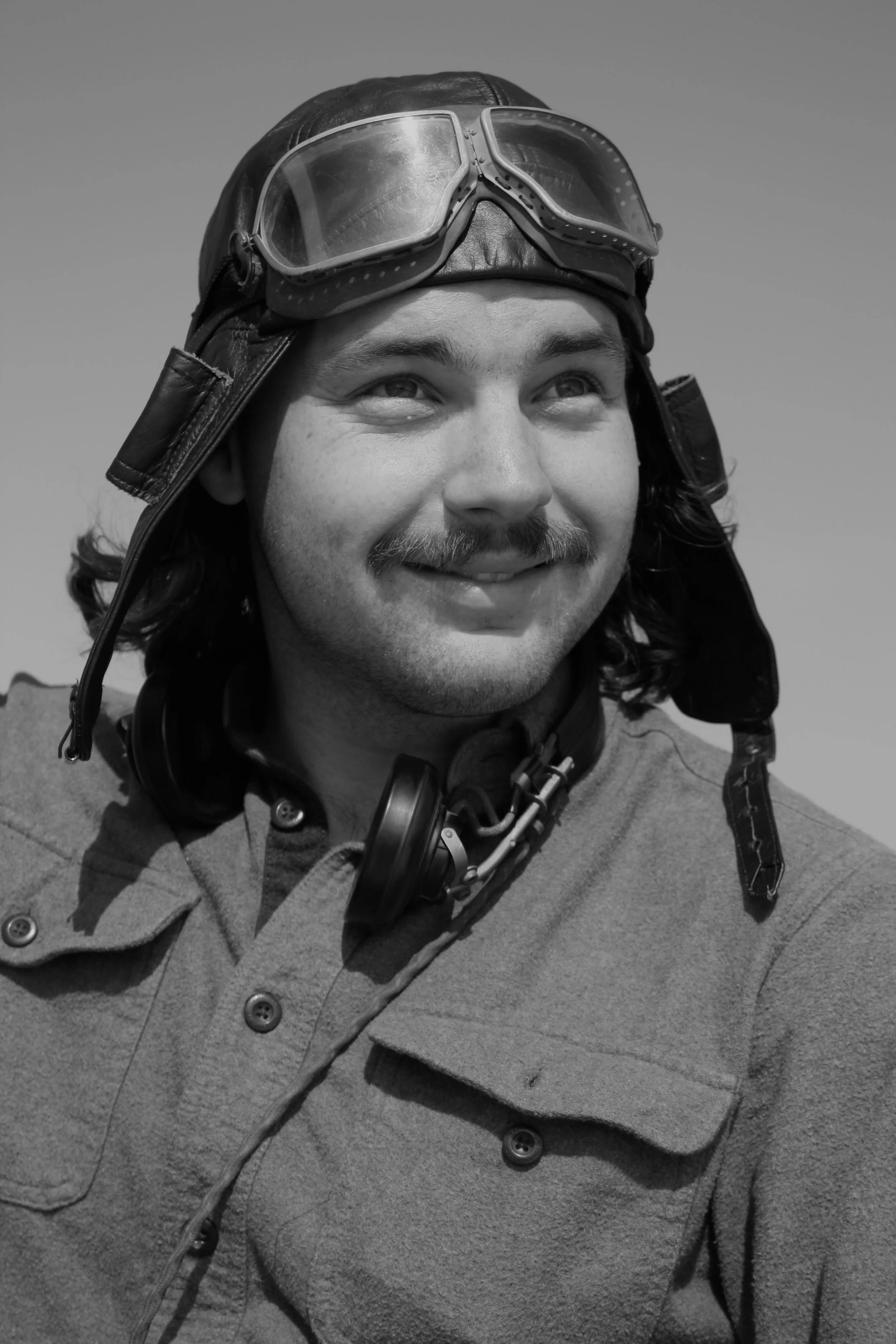 A man wearing aviator goggles, a leather pilot hat, a buttoned shirt, and headphones around his neck, looking upwards with a smile.