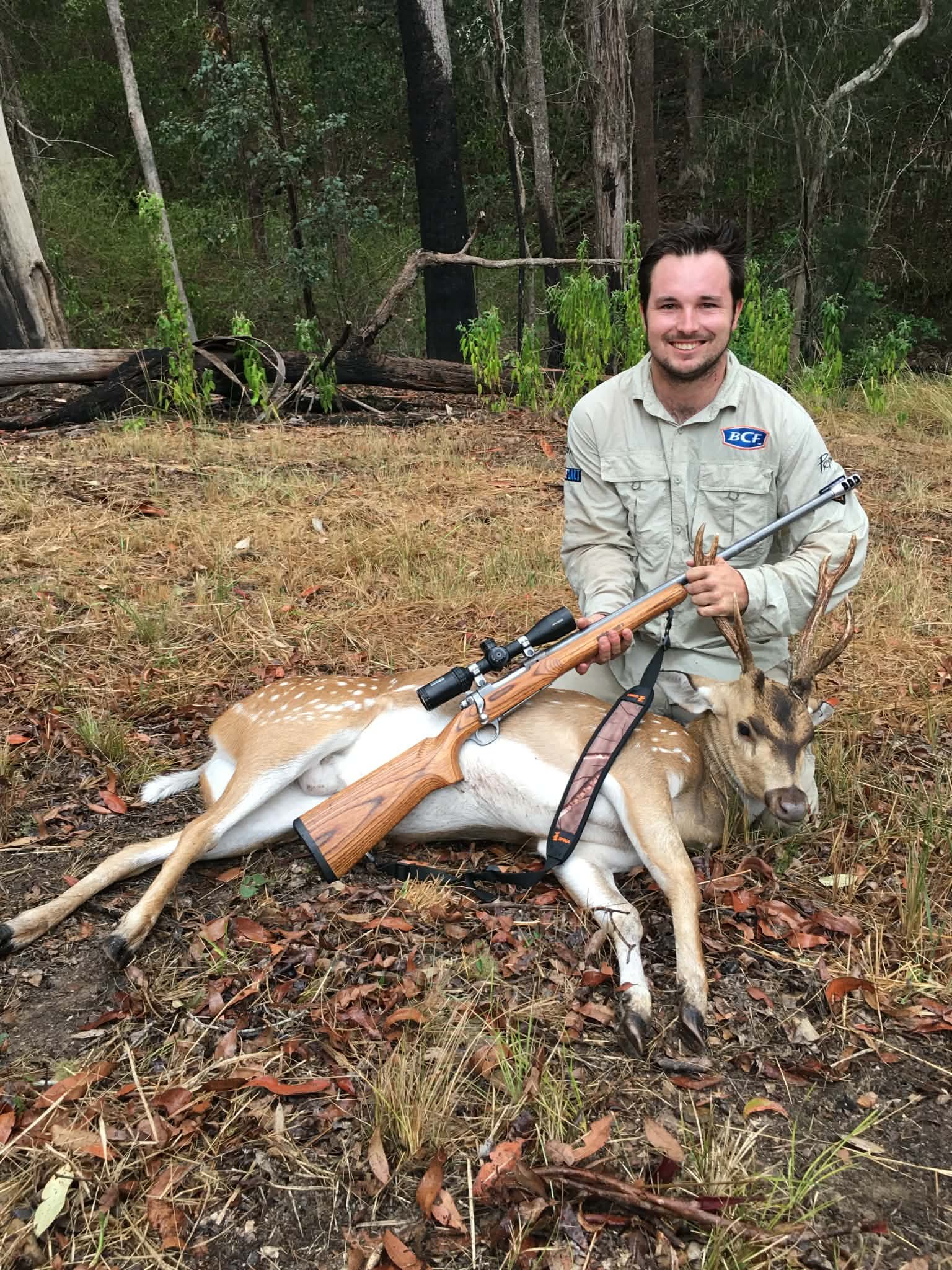 A man sitting on the forest ground with a rifle and a deer in front of him.