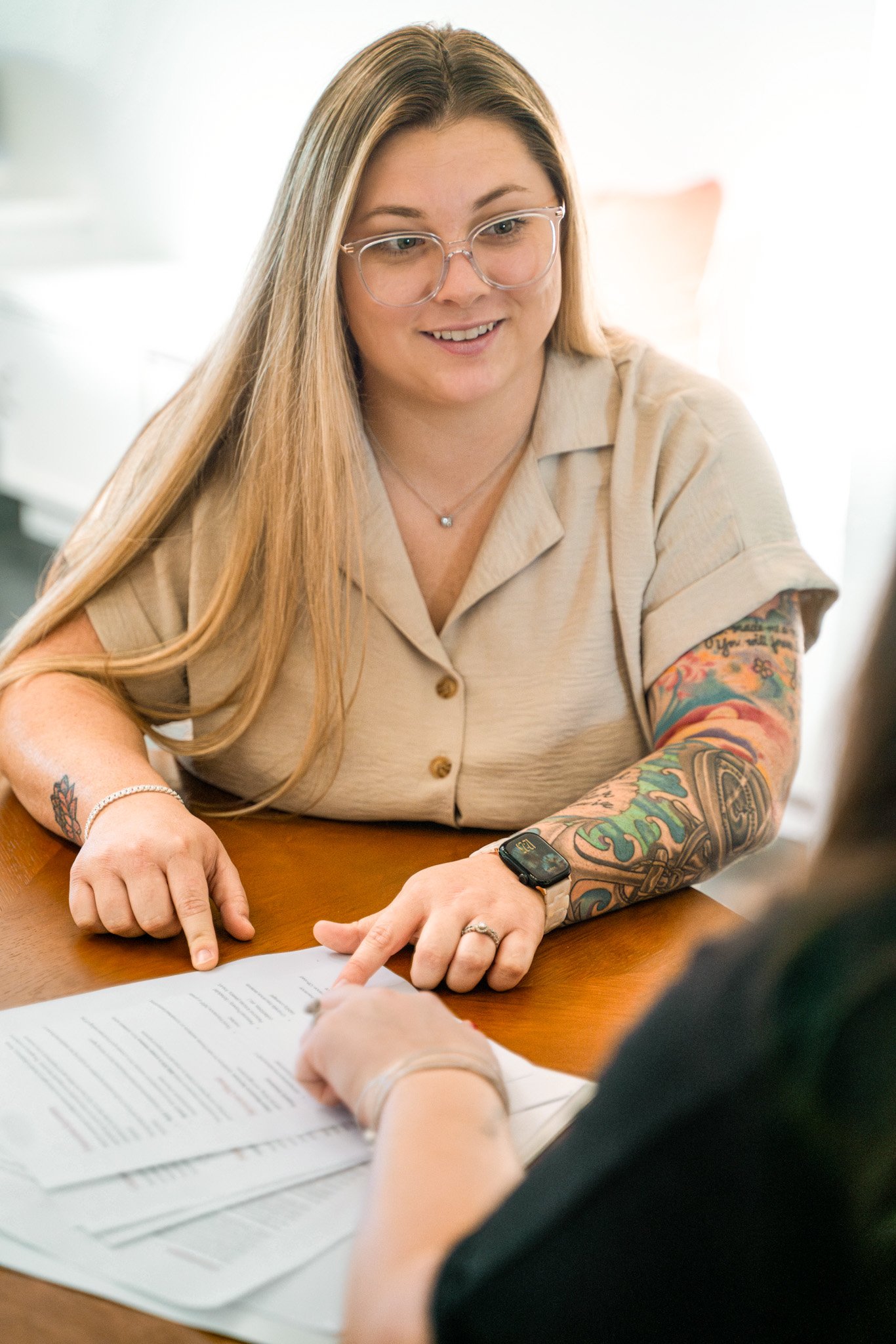Ansley Warner, Florida insurance agent, with long blonde hair, glasses, and colorful tattoos on her arms, smiling and pointing at a document during a discussion with another person at a table.