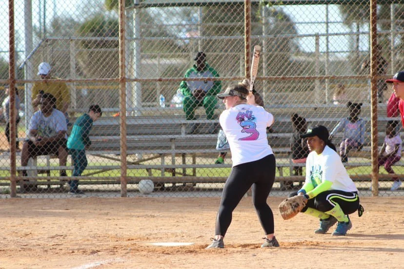 A young woman prepares to hit a softball at a softball game, with other players, a spectator, and a fence in the background.