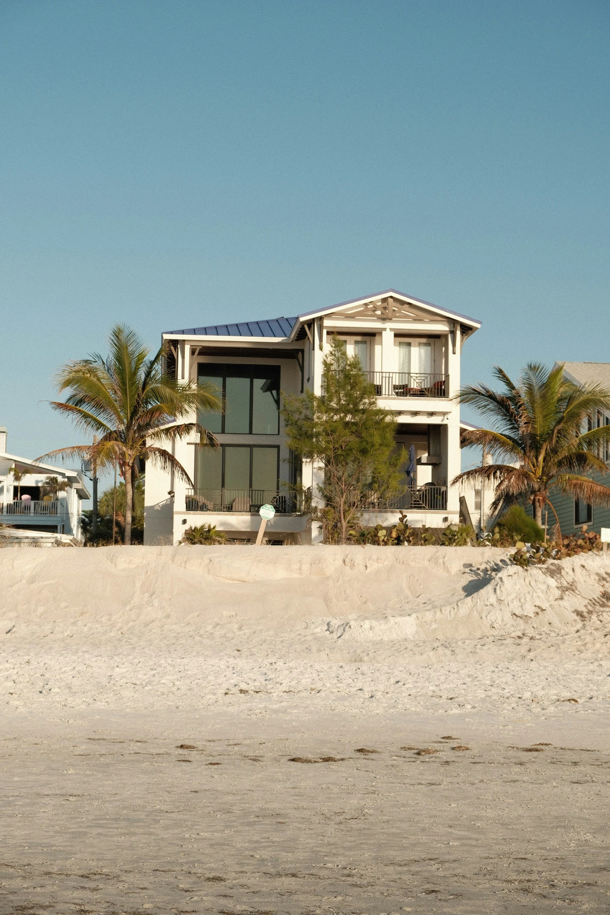 Beach house with large windows, palm trees, and sand in front under a clear blue sky.
