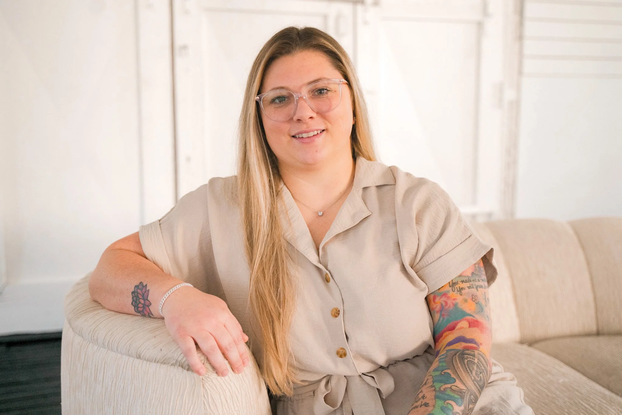 Ansley Warner, Florida insurance agent, pictured with long blonde hair, glasses, and tattoos on her arms sitting on a beige couch and smiling at the camera.