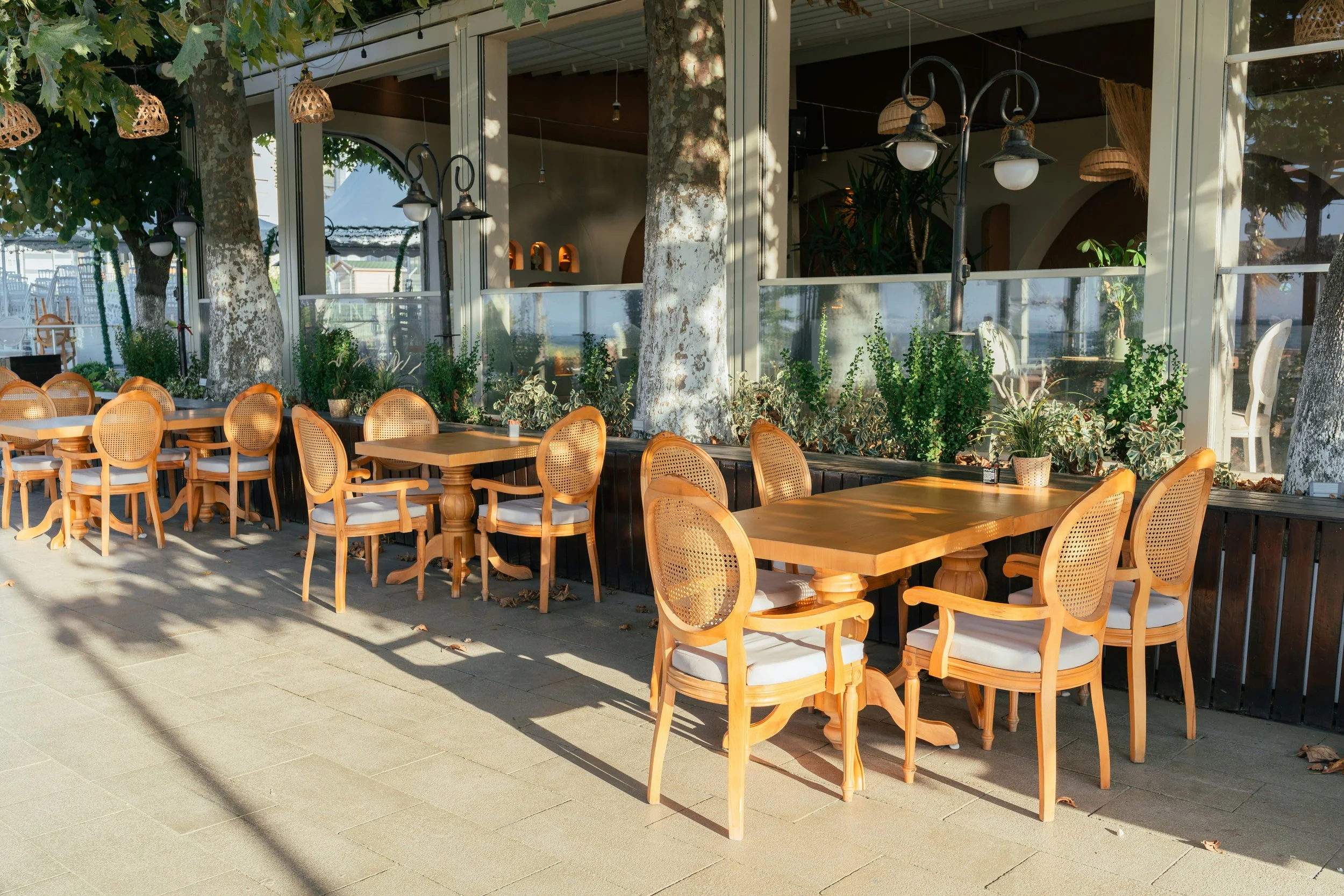 Outdoor seating area with wooden tables and chairs with white cushions, shaded by trees, in front of a glass-walled building with plants inside.