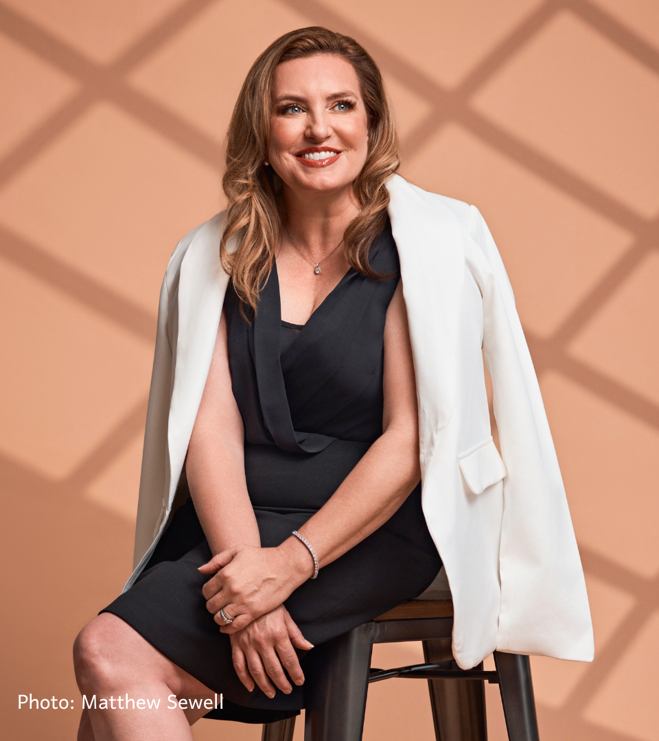 A woman with shoulder-length wavy blonde hair smiling while sitting on a chair. She is wearing a black dress with a white blazer draped over her shoulders and has a necklace and bracelet. The background is peach-colored with a geometric pattern.