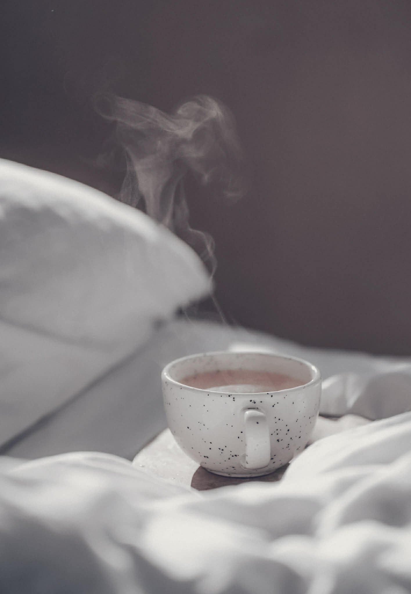 A white ceramic mug with black speckles, filled with hot beverage, sitting on white bedding with steam rising from it. The background is dark and blurred.