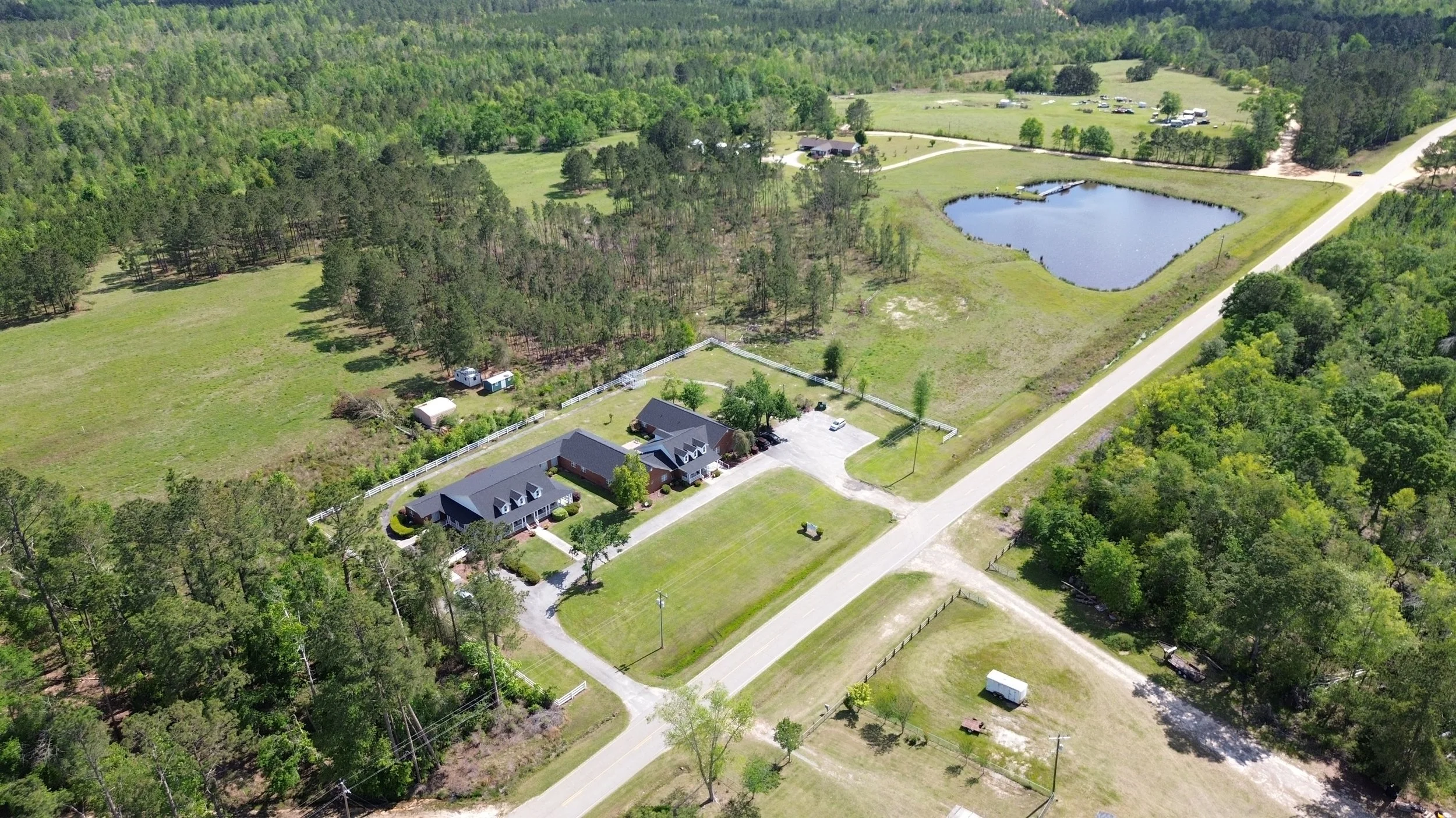 Aerial view of a rural property with a house, detached garage, fenced yard, pond, and surrounding greenery and wooded areas.