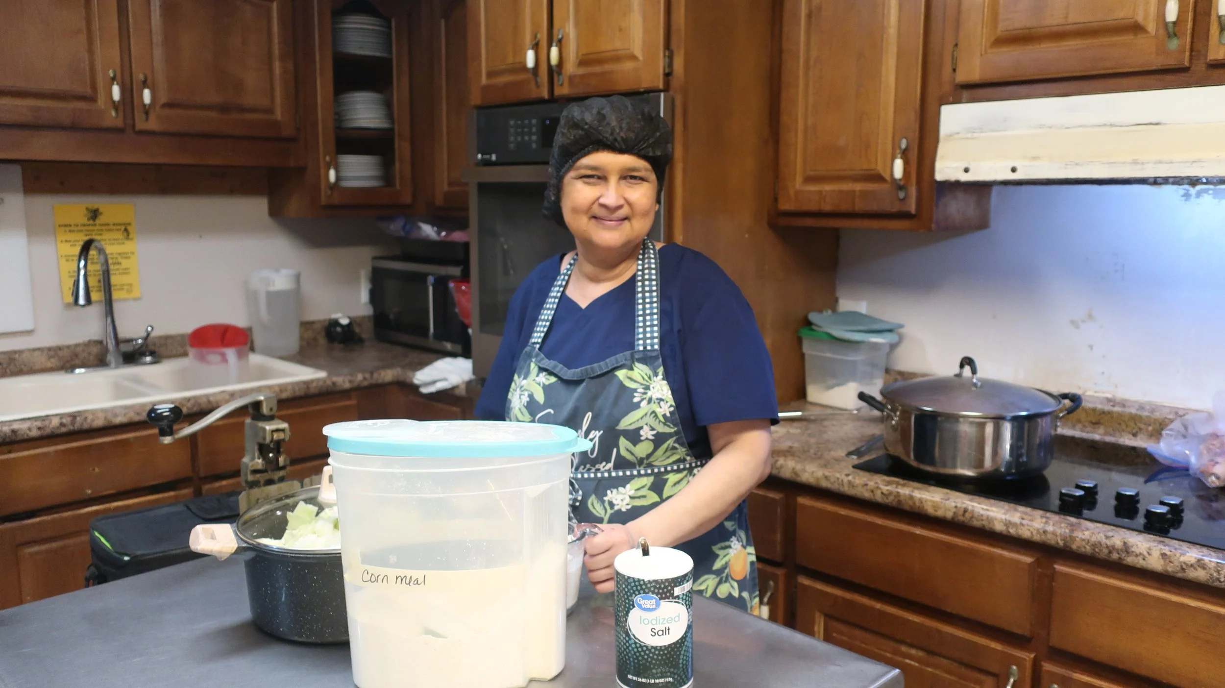 A smiling woman in a kitchen wearing a black hair cover and a blue apron with a floral pattern, standing at a countertop with cooking ingredients and utensils.