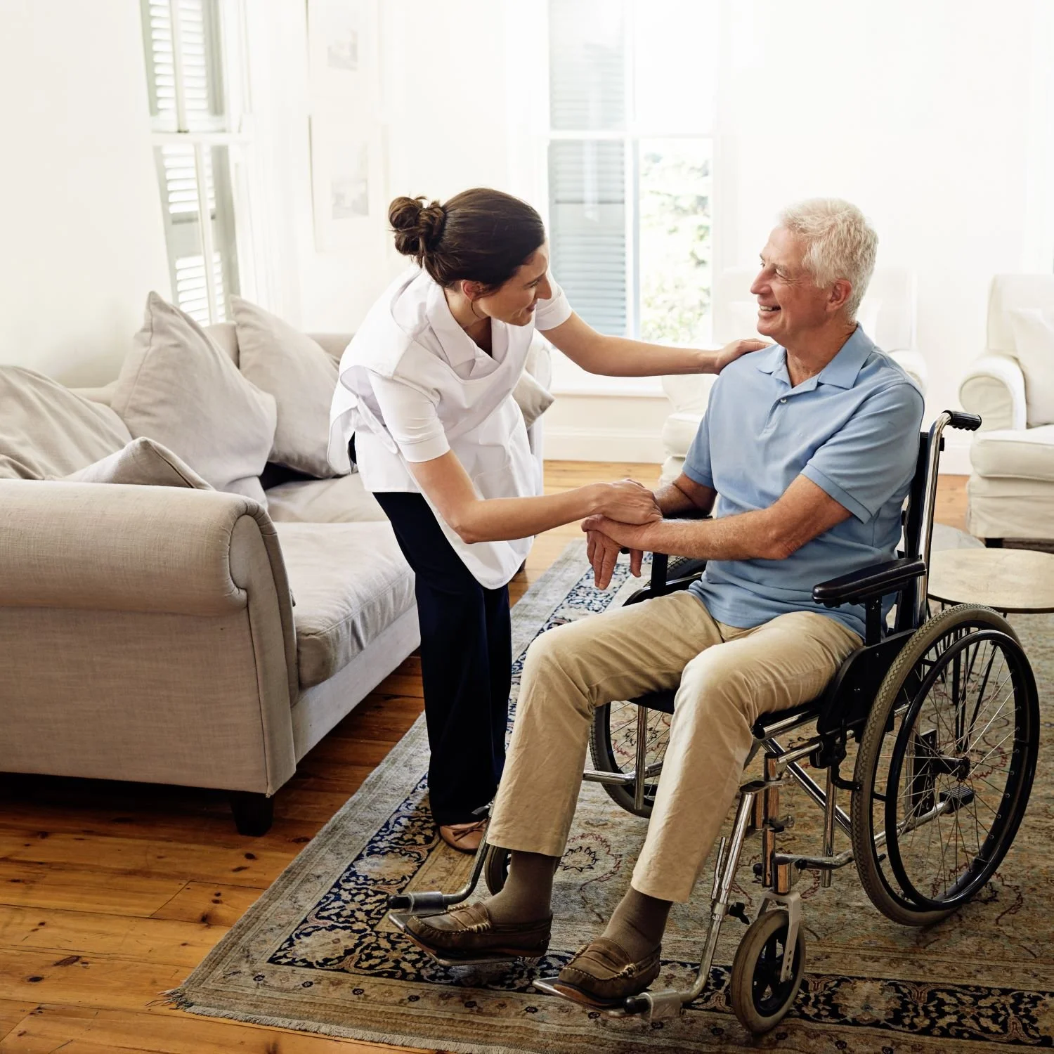 A nurse talking to an elderly man in a wheelchair in a bright living room.