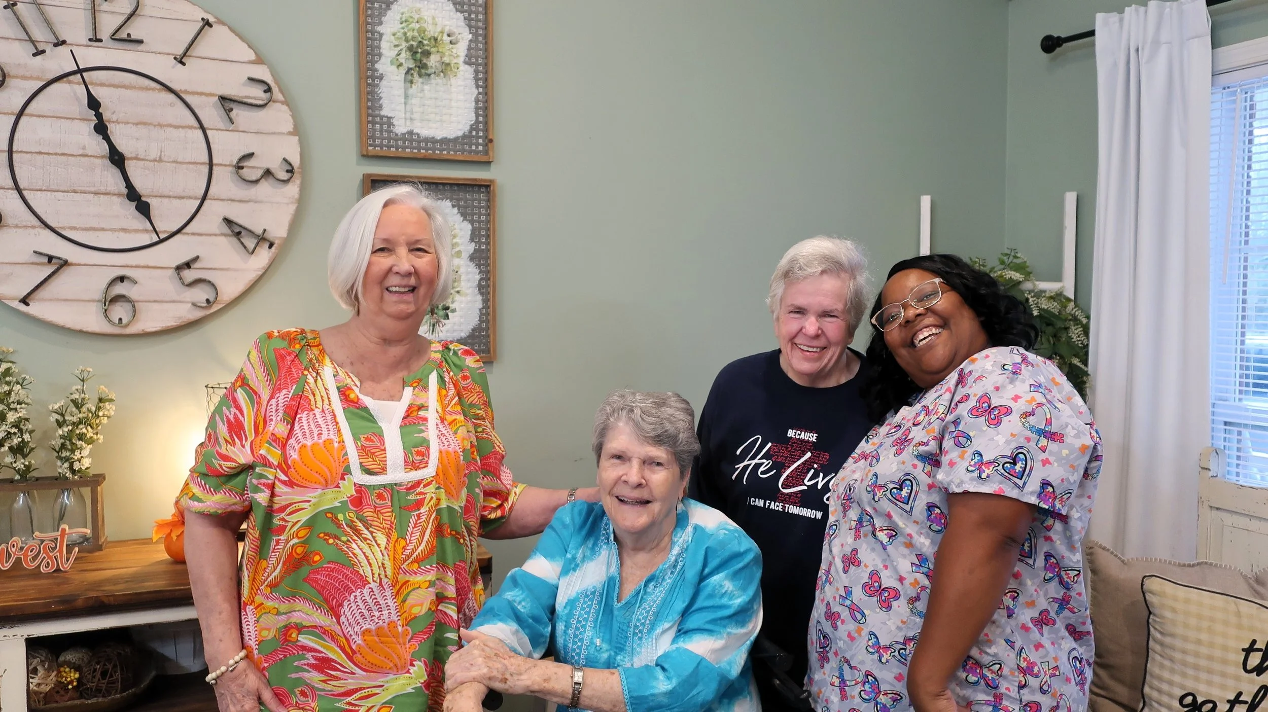Four women and one elderly woman gathered together in a living room, smiling and enjoying each other's company. The women are wearing colorful tops, with one woman in scrubs, and the elderly woman is seated in a wheelchair. Behind them, there is a large wall clock and some wall art, with curtains and potted plants in the background.
