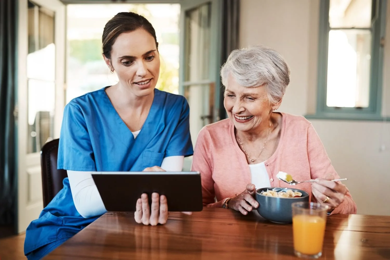 A nurse showing something on a tablet to an elderly woman eating cereal at a wooden table with a glass of orange juice nearby.