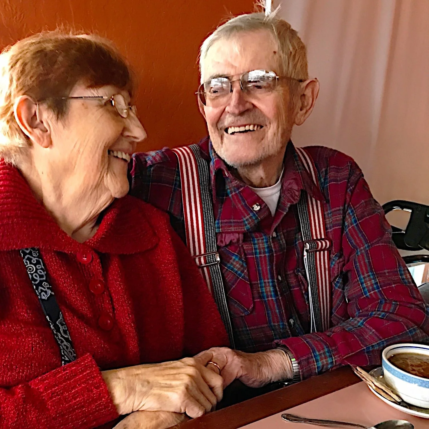 An elderly couple sitting at a table, smiling and holding hands, with a bowl of soup in front of them, sharing a joyful moment.