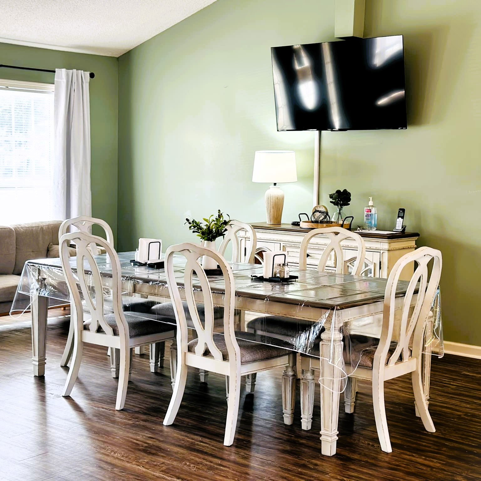Dining room with six white chairs around a table, a large TV on the wall, a sideboard with a lamp and decor, and a window with white curtains.