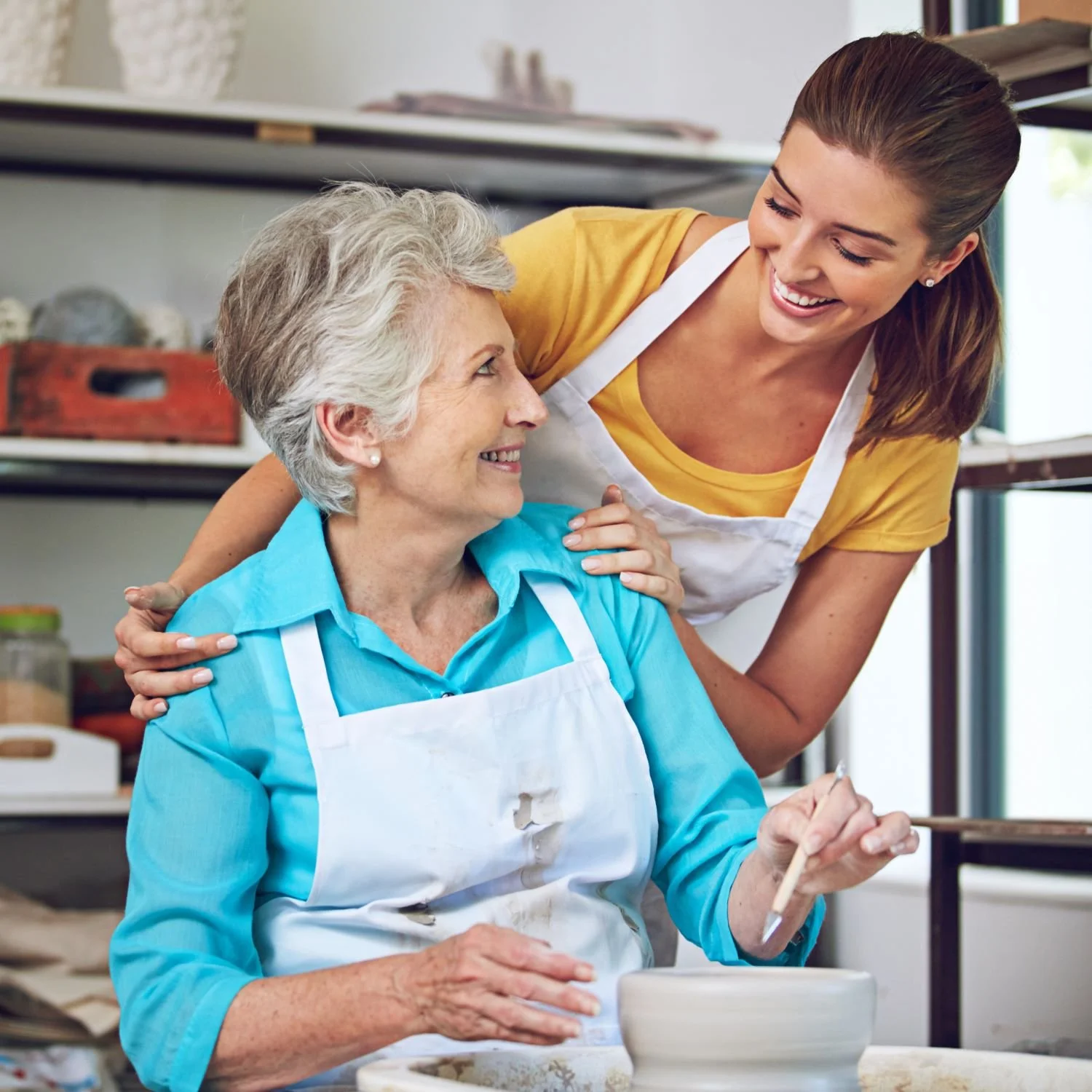A young woman helps an elderly woman paint pottery in a studio.