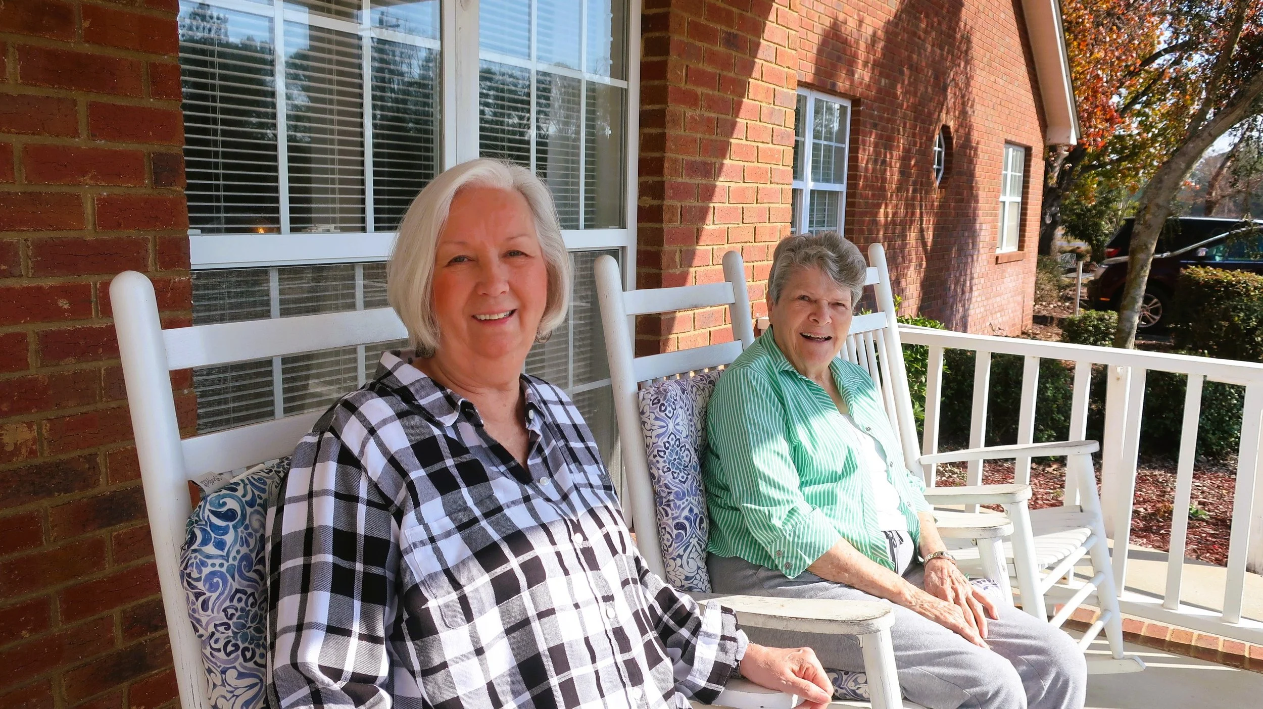 Two elderly women sitting on white porch rocking chairs on a sunny day, smiling, with a brick house and trees in the background.