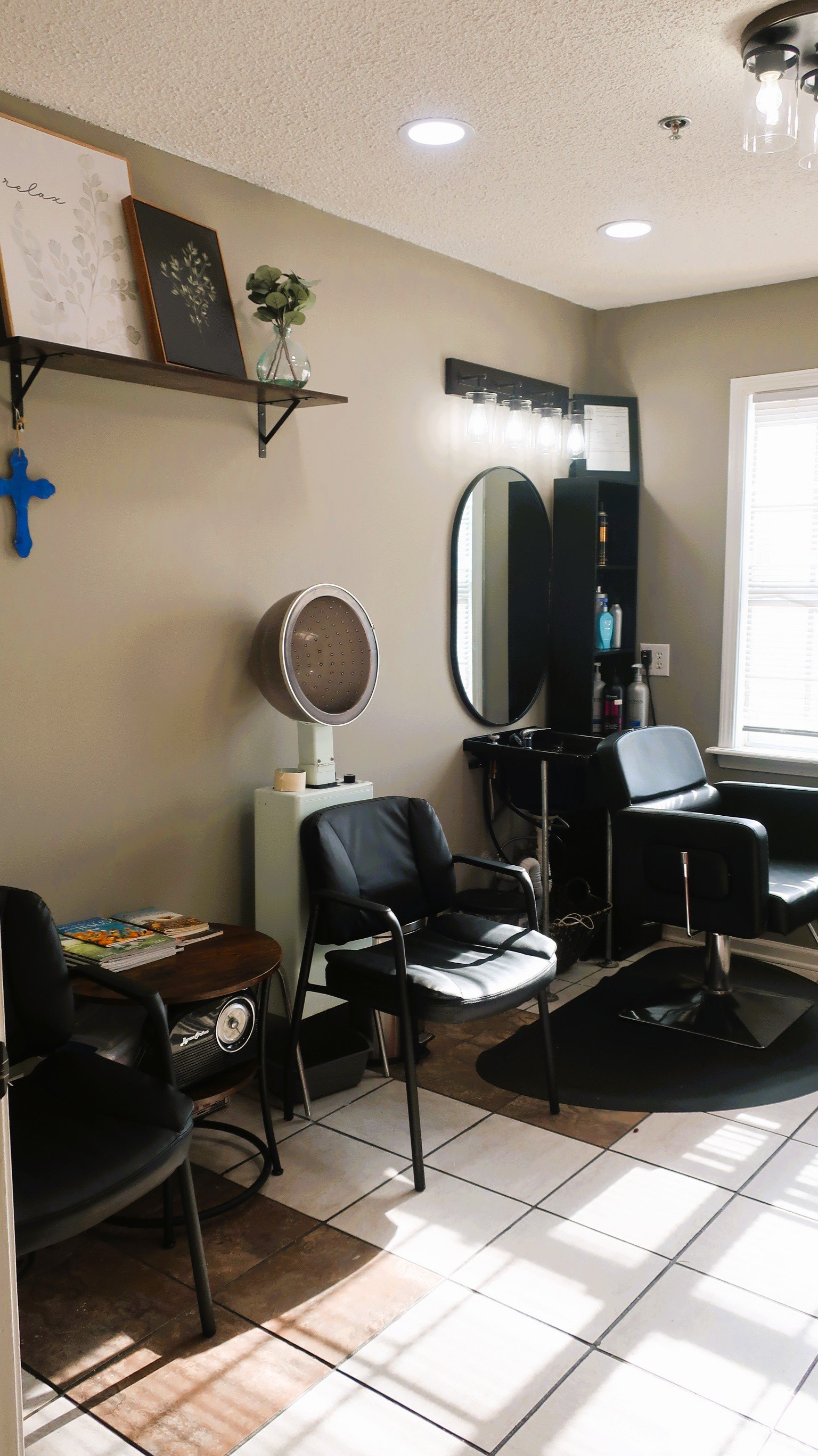 Interior of a hair salon with black styling chairs, a mirror, shelves with hair products, a hair dryer, and decorative items on the wall.